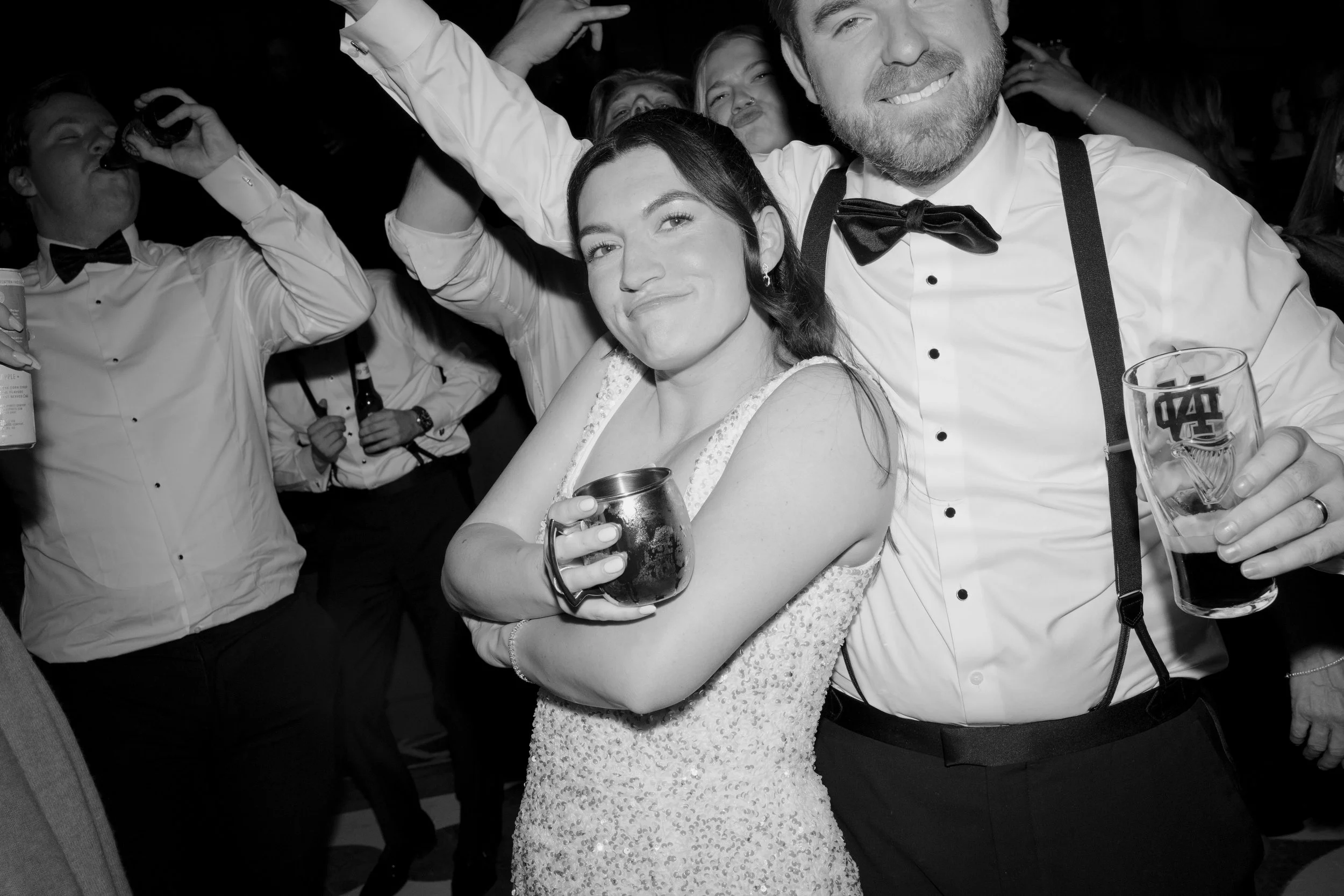 A group of people at a party or celebration, with a woman in a white dress holding a drink and smiling, and a man in a tuxedo with suspenders smiling as well. Others are in the background, some holding drinks and enjoying the event.