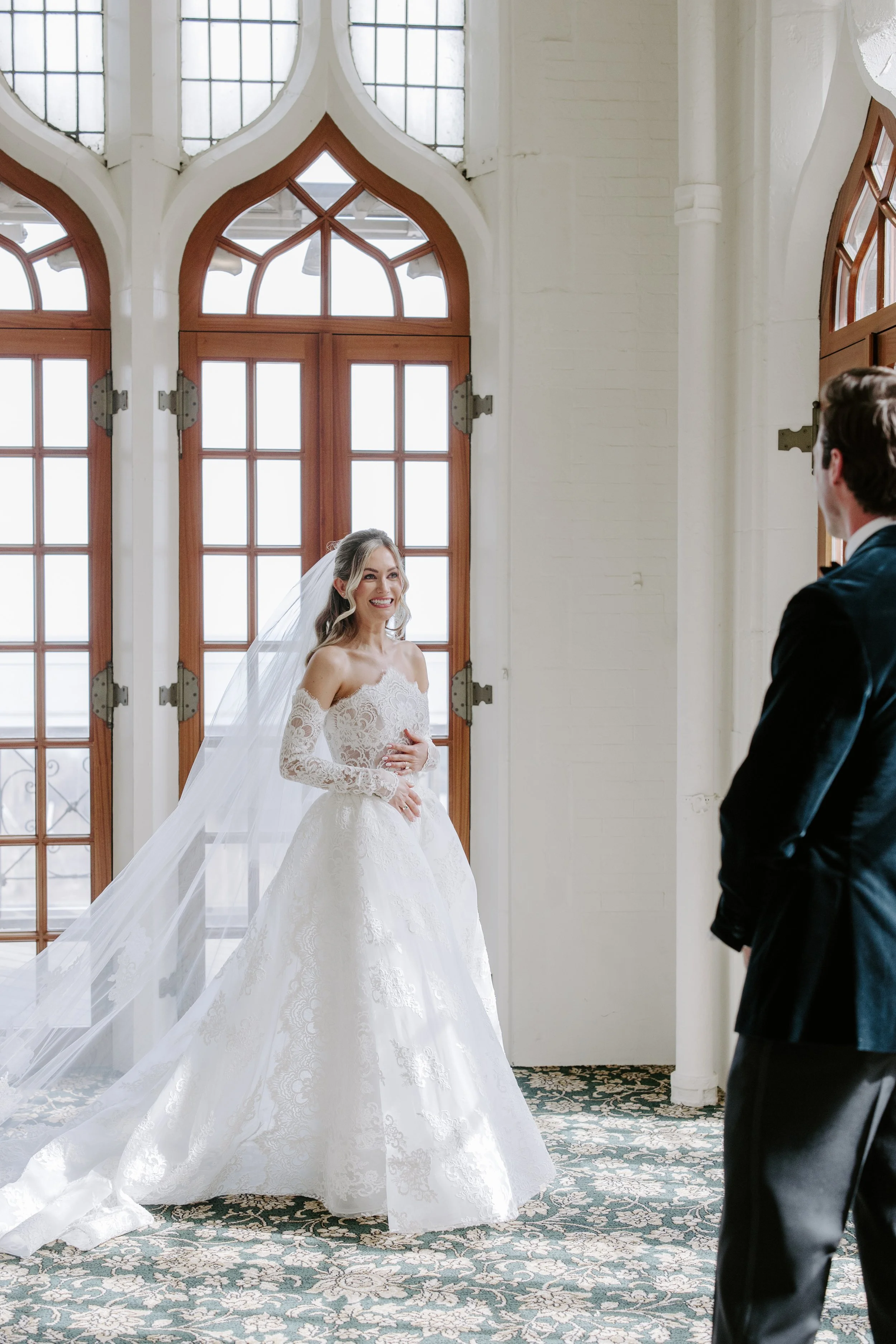 A bride in a white lace wedding dress with long sleeves and a veil, smiling and holding her stomach, standing in front of large windows. A groom in a dark suit is facing her, visible from the back.
