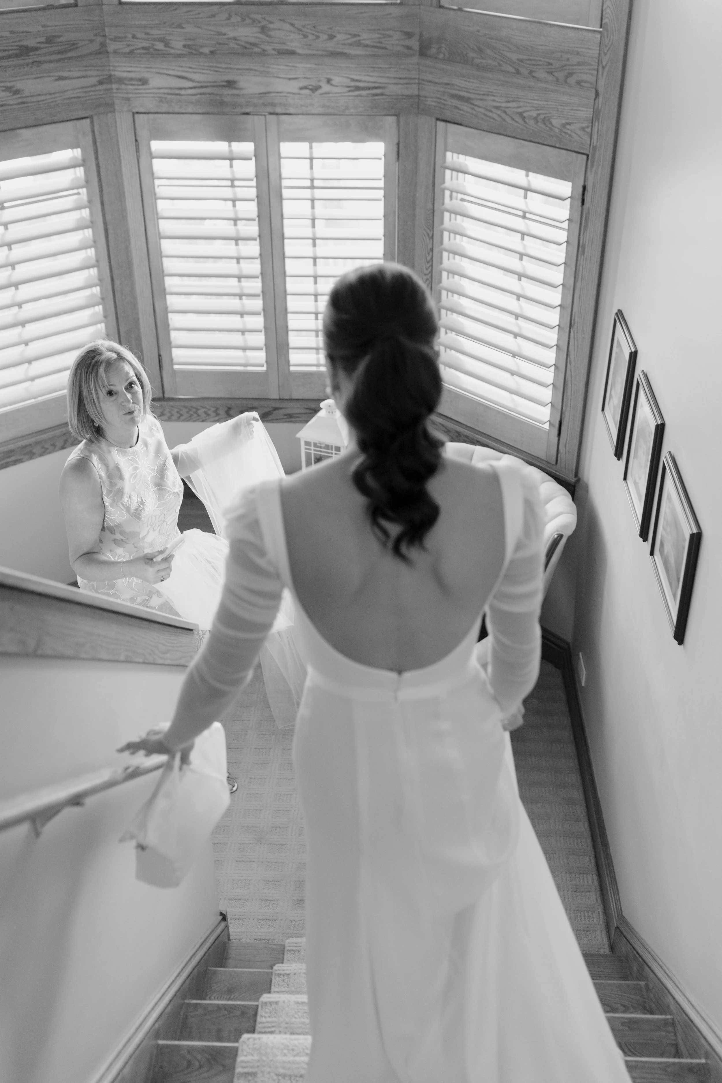 A woman in a white wedding dress looking into a mirror, with a young woman in a white outfit standing behind her. The scene is in a room with large windows with blinds and wooden trim.