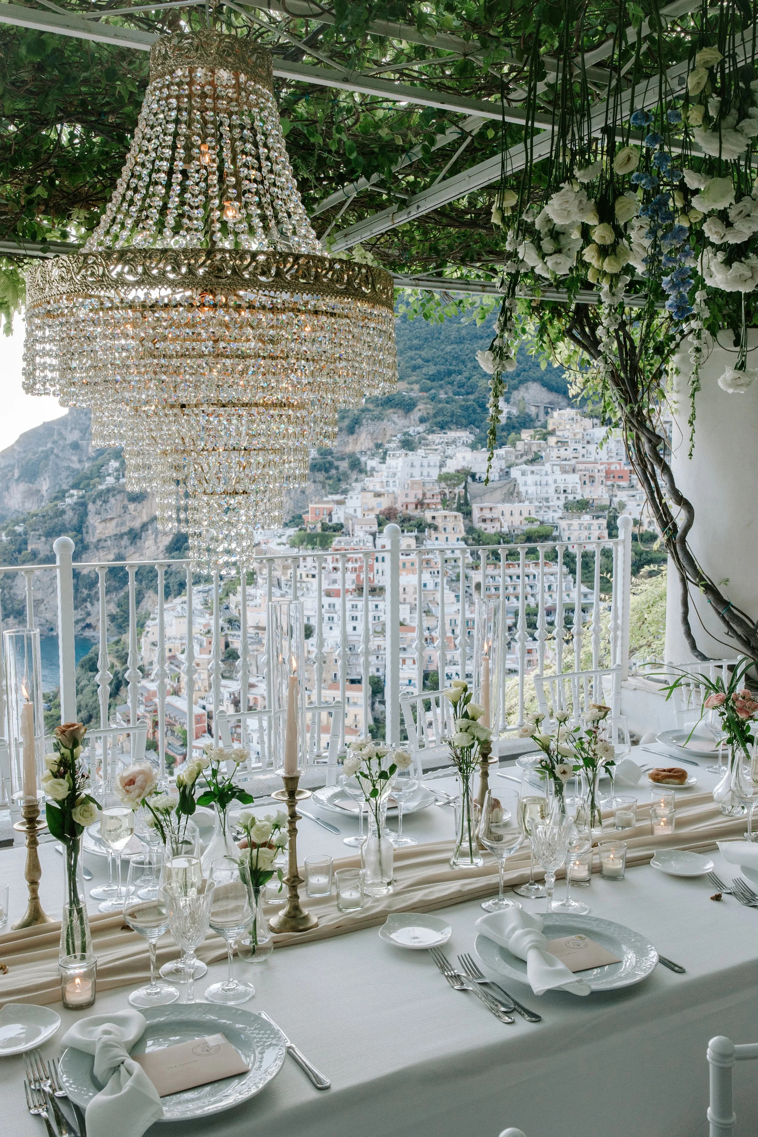 Elegant dining setup on a terrace with white tablecloths, crystal chandeliers, floral centerpieces, and a scenic hillside view of white buildings.