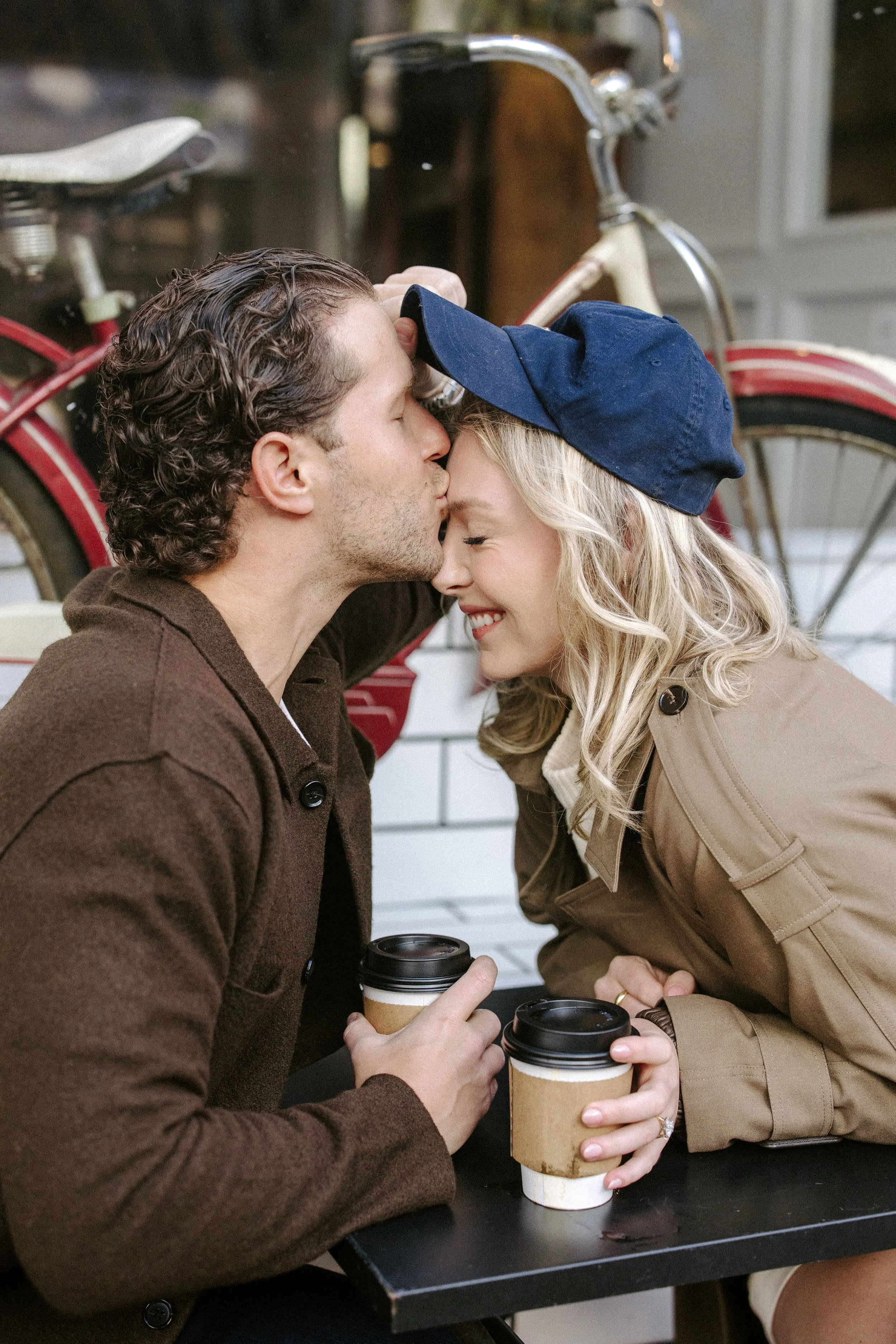A man and woman are sitting at a table outside, sharing a tender moment with their foreheads touching and eyes closed. They are holding coffee cups and wearing casual coats, with the man wearing a brown coat and the woman a beige trench coat and a na