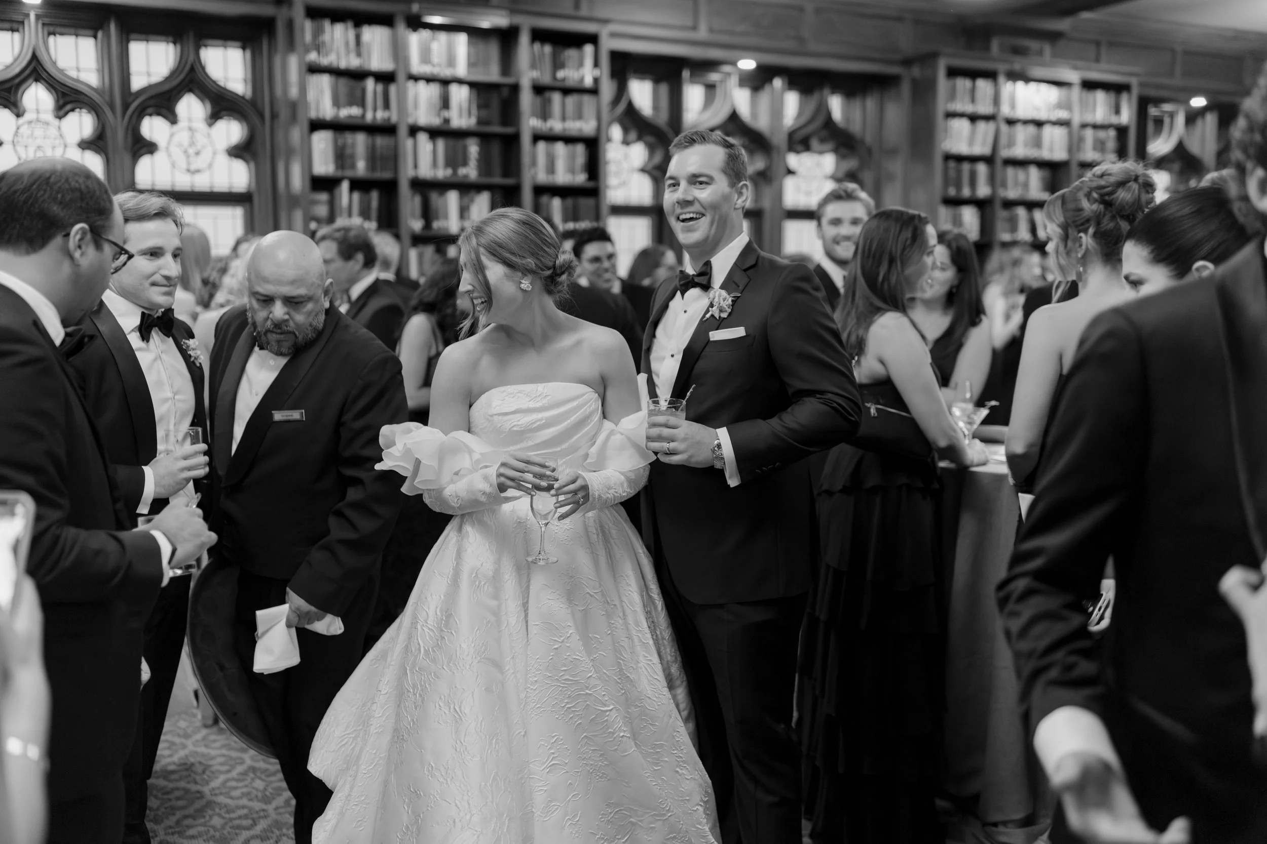 Black and white photo of a wedding reception with guests in formal attire inside a library or study room with tall bookshelves and stained glass windows. A bride in a strapless wedding gown and a groom in a tuxedo are smiling, holding glasses, and su
