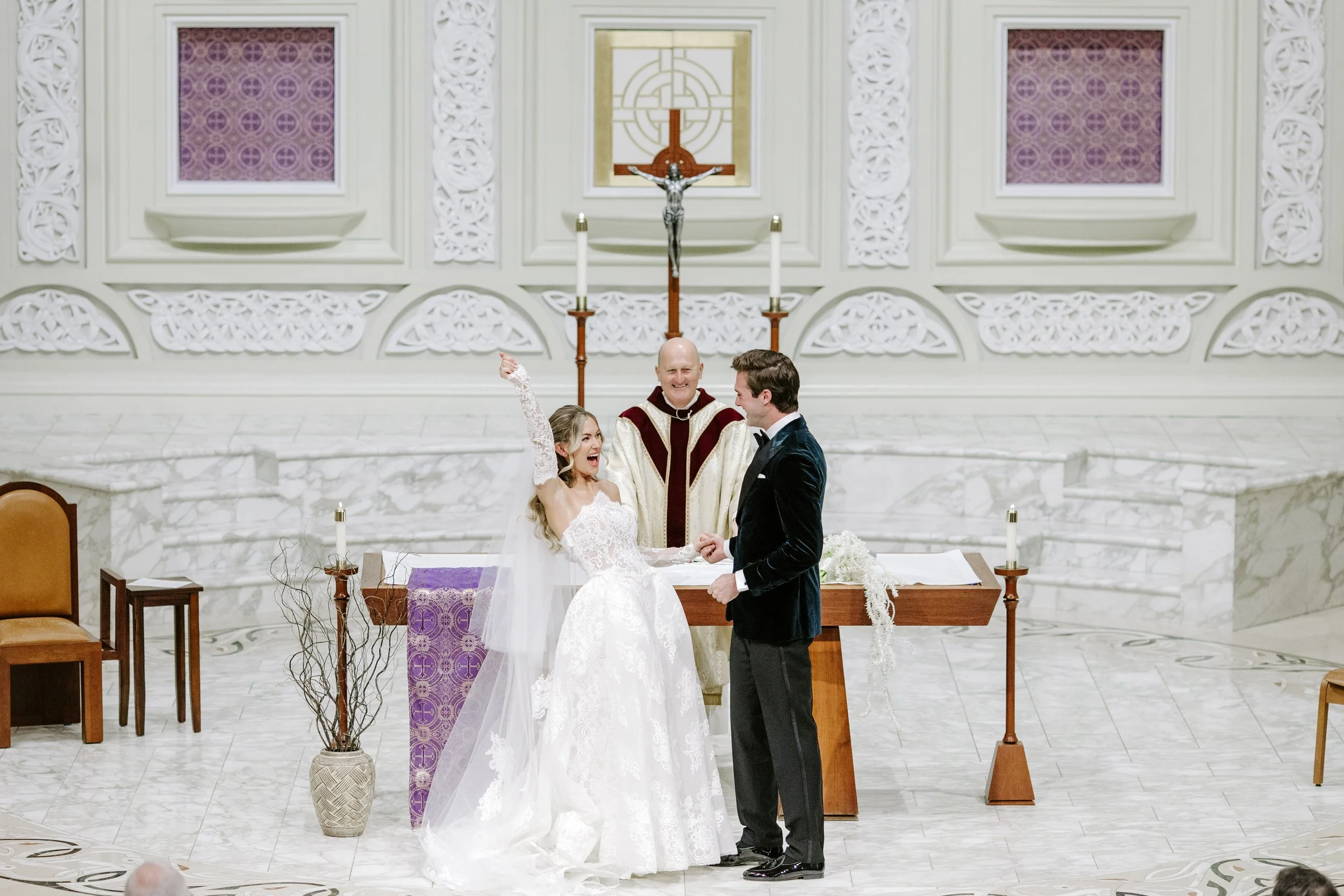 A bride and groom exchanging rings during their wedding ceremony inside a church, with a priest officiating. The bride is in a white lace gown, and the groom is in a dark tuxedo. The church has white ornate decorations, candles, and religious symbols