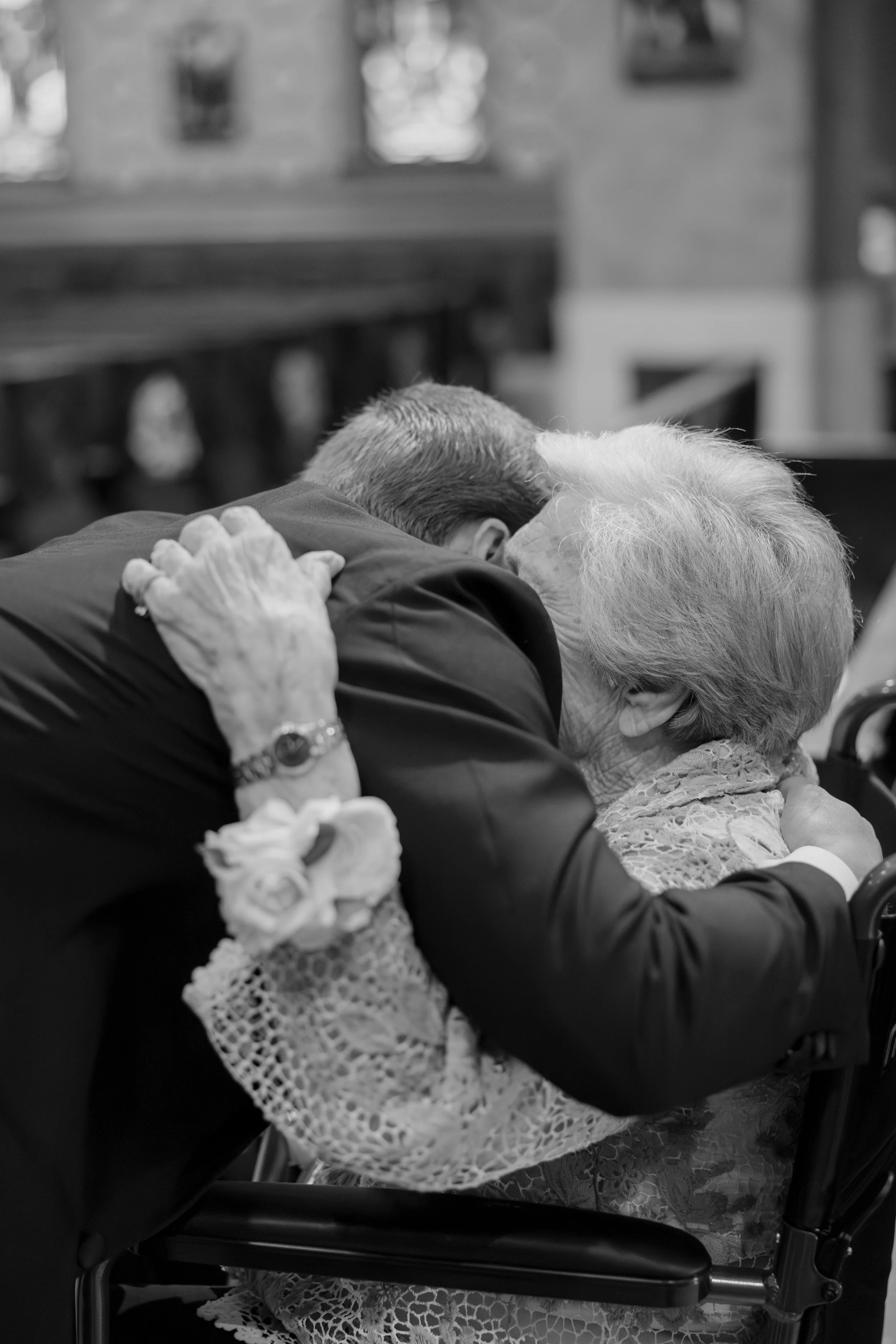 An elderly woman with white hair and a lace dress is being kissed by a man in a dark suit at a formal event or gathering. The man has his arm around her, and they are seated in a wheelchair.