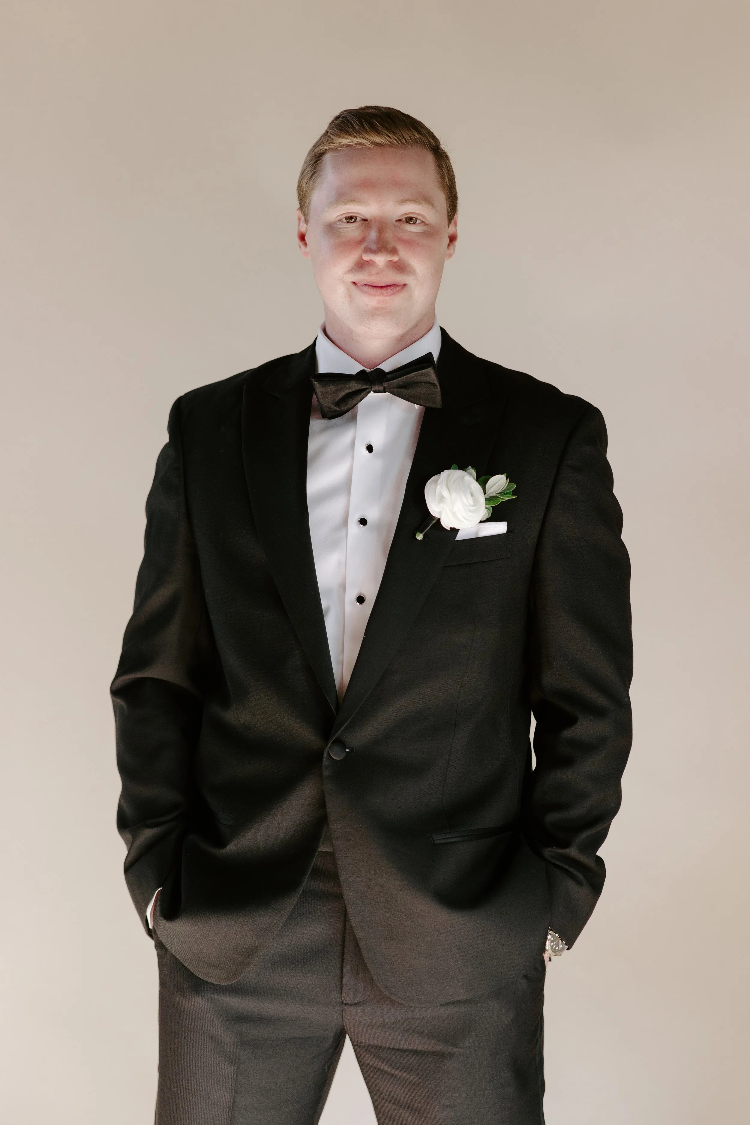 A young man wearing a black tuxedo with a white dress shirt, black bow tie, and boutonniere, posing against a plain neutral background.