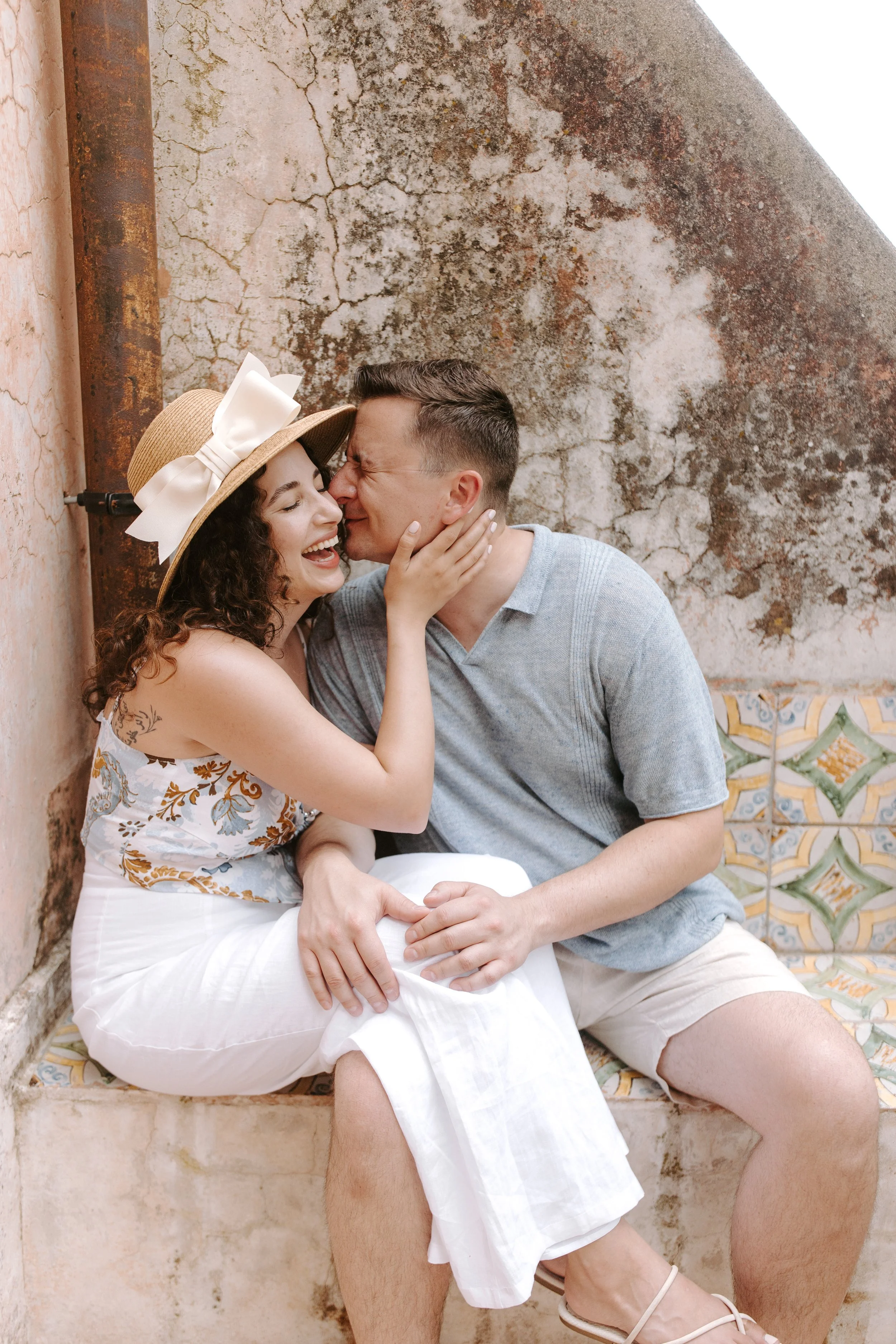 A couple sitting close together on a patterned tiled bench with a weathered, cracked wall in the background. The woman is wearing a straw hat with a large white bow, has curly hair, and is smiling while touching the man's face. The man is wearing a l