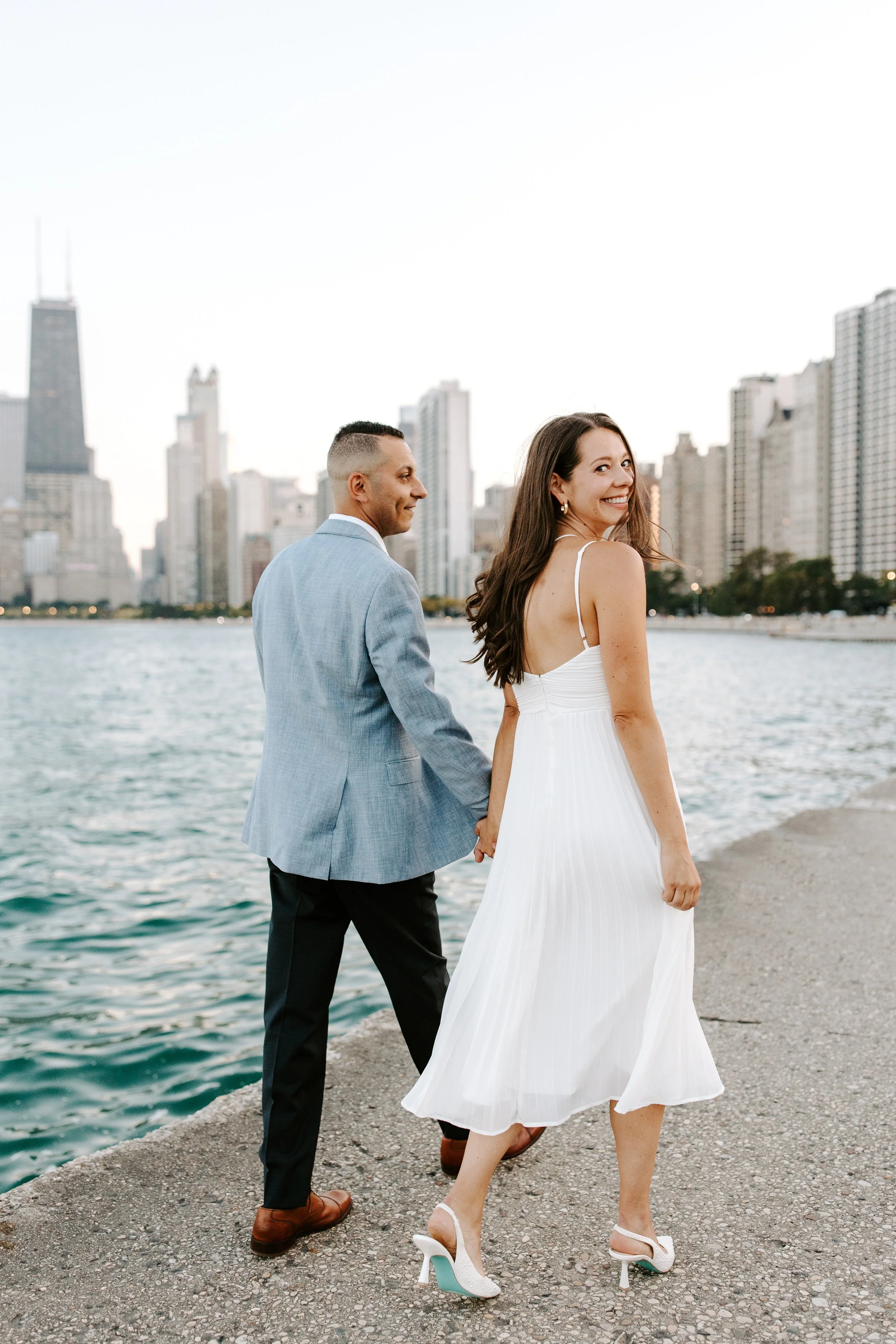 A smiling woman wearing a white dress and high heels is holding hands with a man dressed in a light blue blazer and dark pants along a shoreline with water and city skyline in the background.