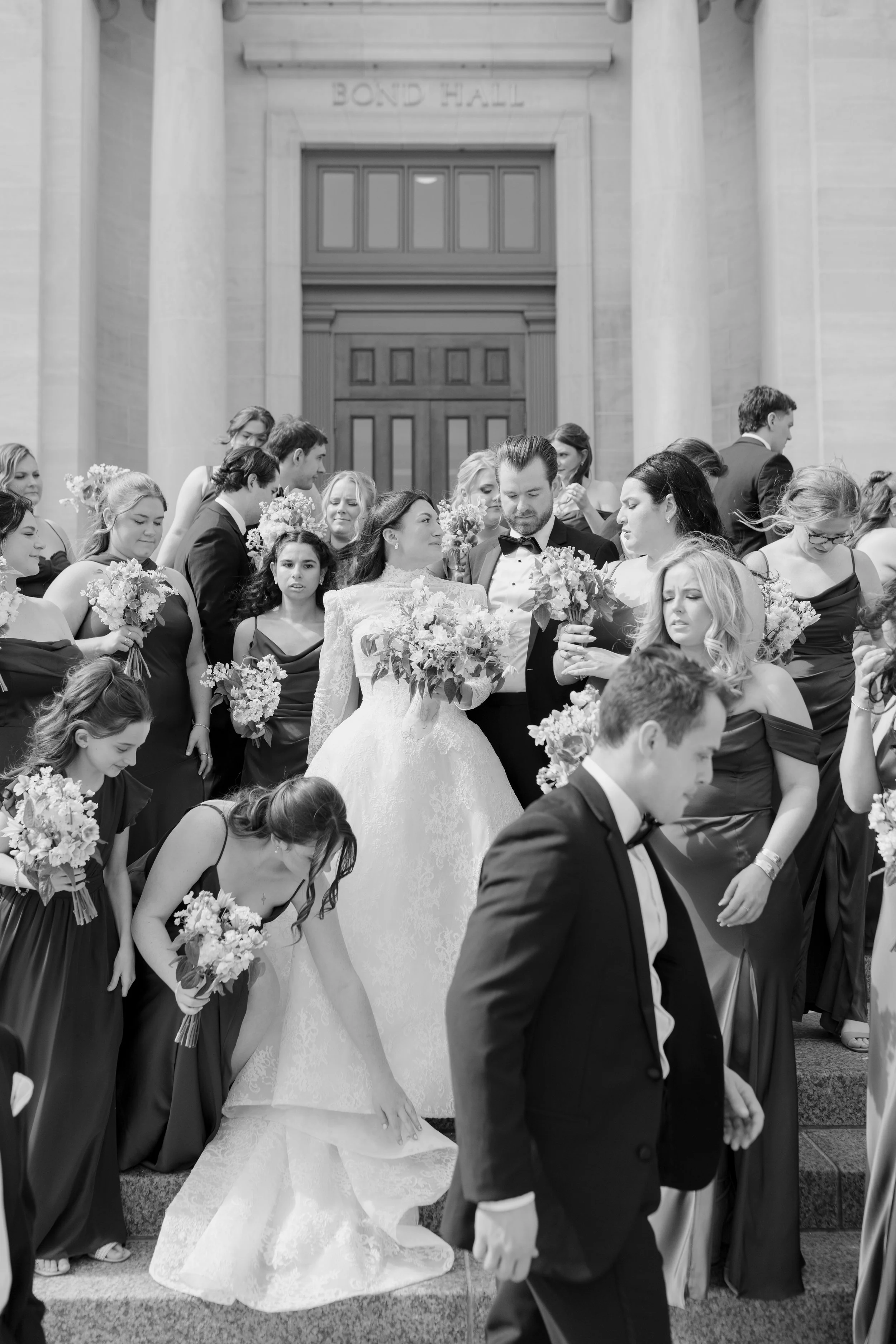 A wedding group exiting a building labeled 'BOND HALL' with several bridesmaids and groomsmen, including a bride in a white gown and a groom in a tuxedo, holding bouquets of flowers.