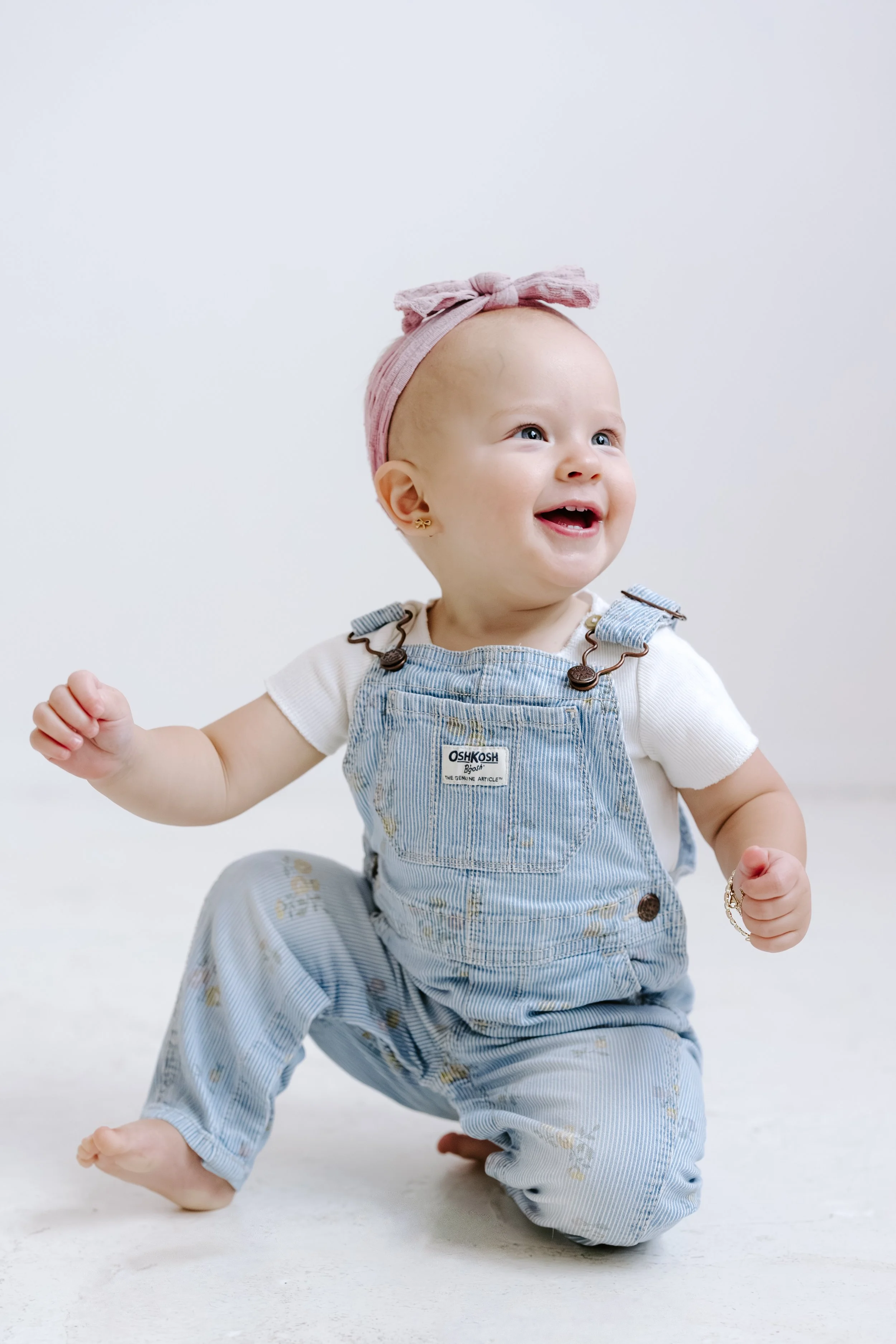 Smiling baby girl with a light pink headband and gold earrings, sitting on the floor in overalls and a white shirt, looking happy and energetic.