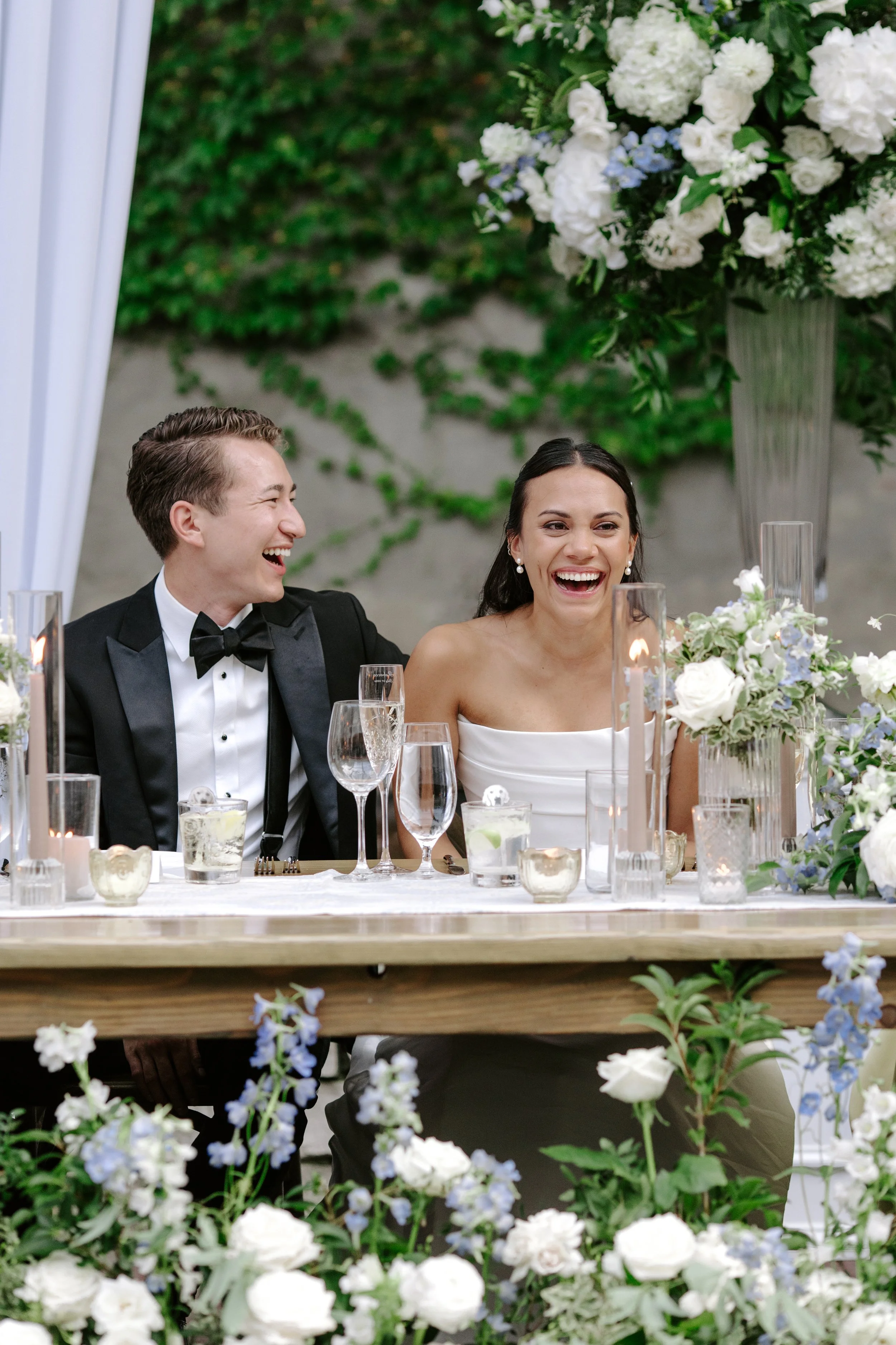Two people, a man in a tuxedo and a woman in a white dress, smiling and laughing at a wedding reception table decorated with flowers, candles, and glassware.