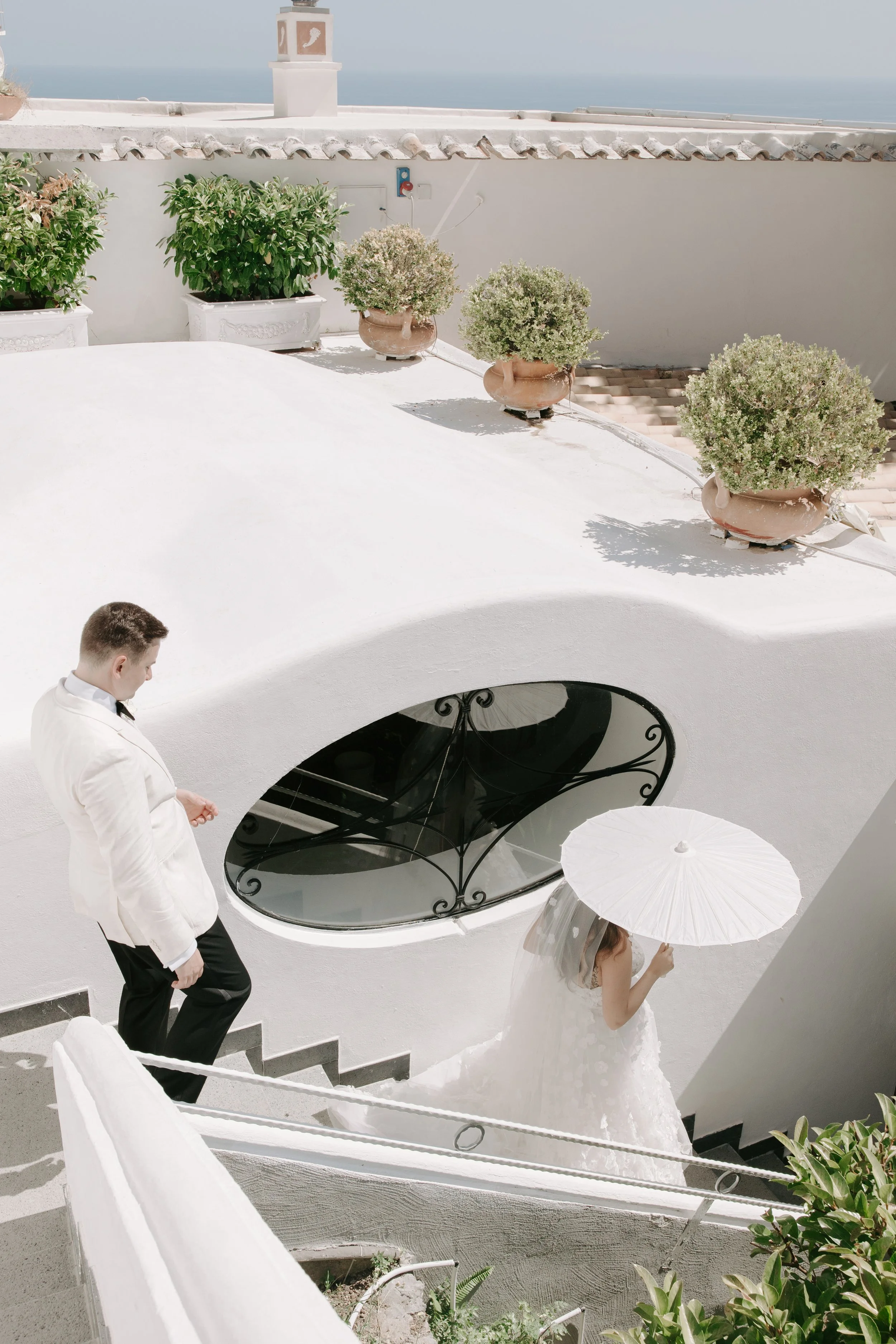 A wedding scene on a white terrace with potted plants, a brief glimpse of a bride holding a white parasol, and a man dressed in a white jacket and black pants looking downward, with a view of the ocean in the background.