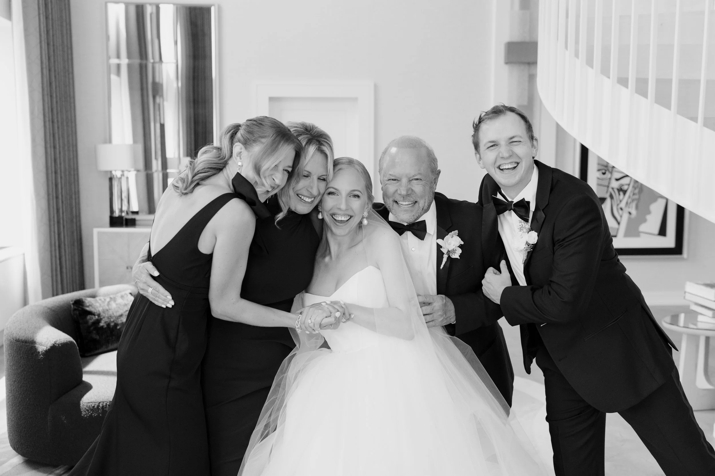 A group of six people, including a bride in a wedding gown and five others in formal attire, laughing and hugging in a modern indoor setting.
