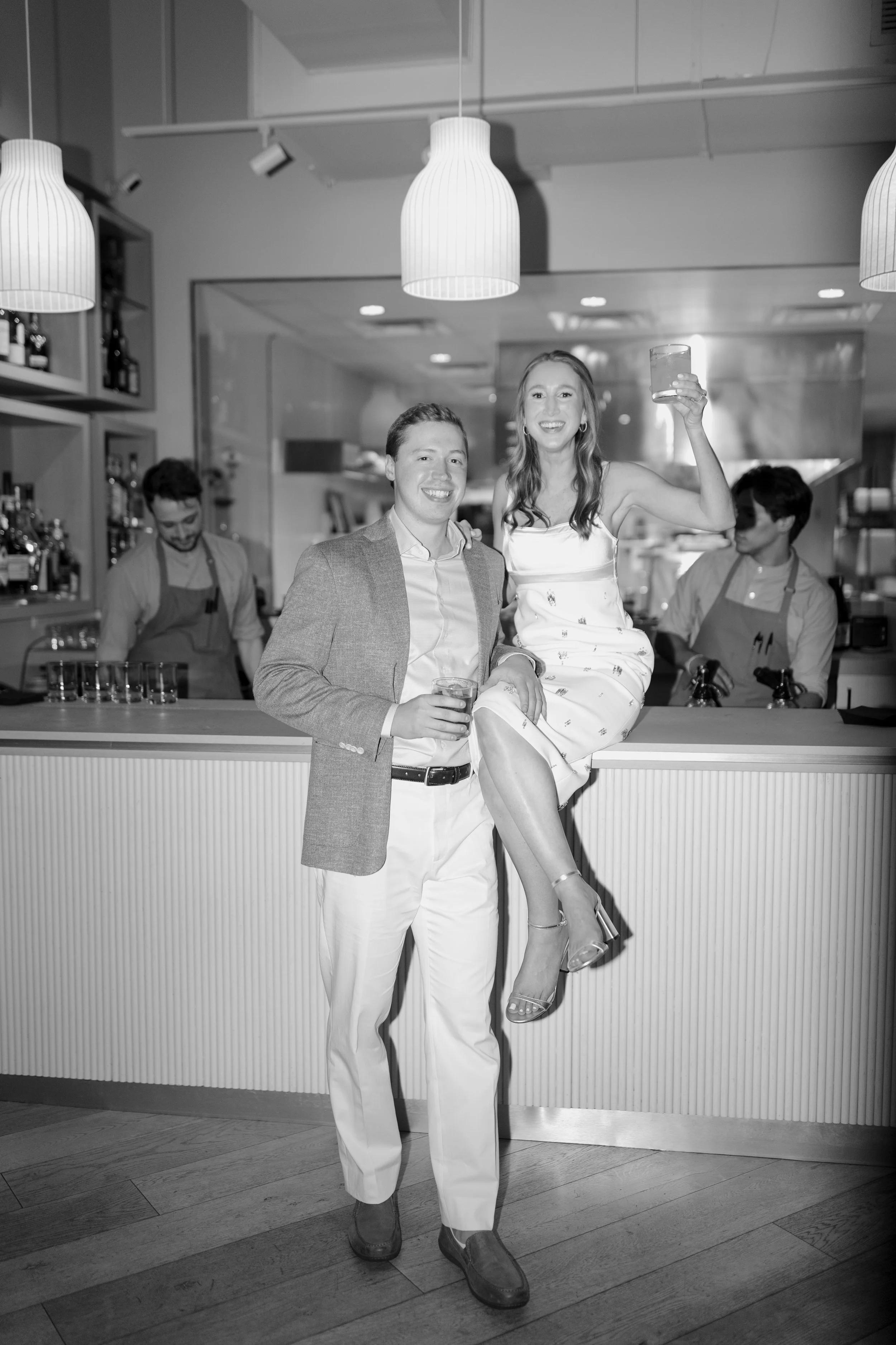 A black and white photo of a man and a woman at a bar. The man is wearing a suit jacket and white pants, holding a drink in his hand, and smiling at the camera. The woman is sitting on the bar counter beside him, raising her drink in a toast, wearing