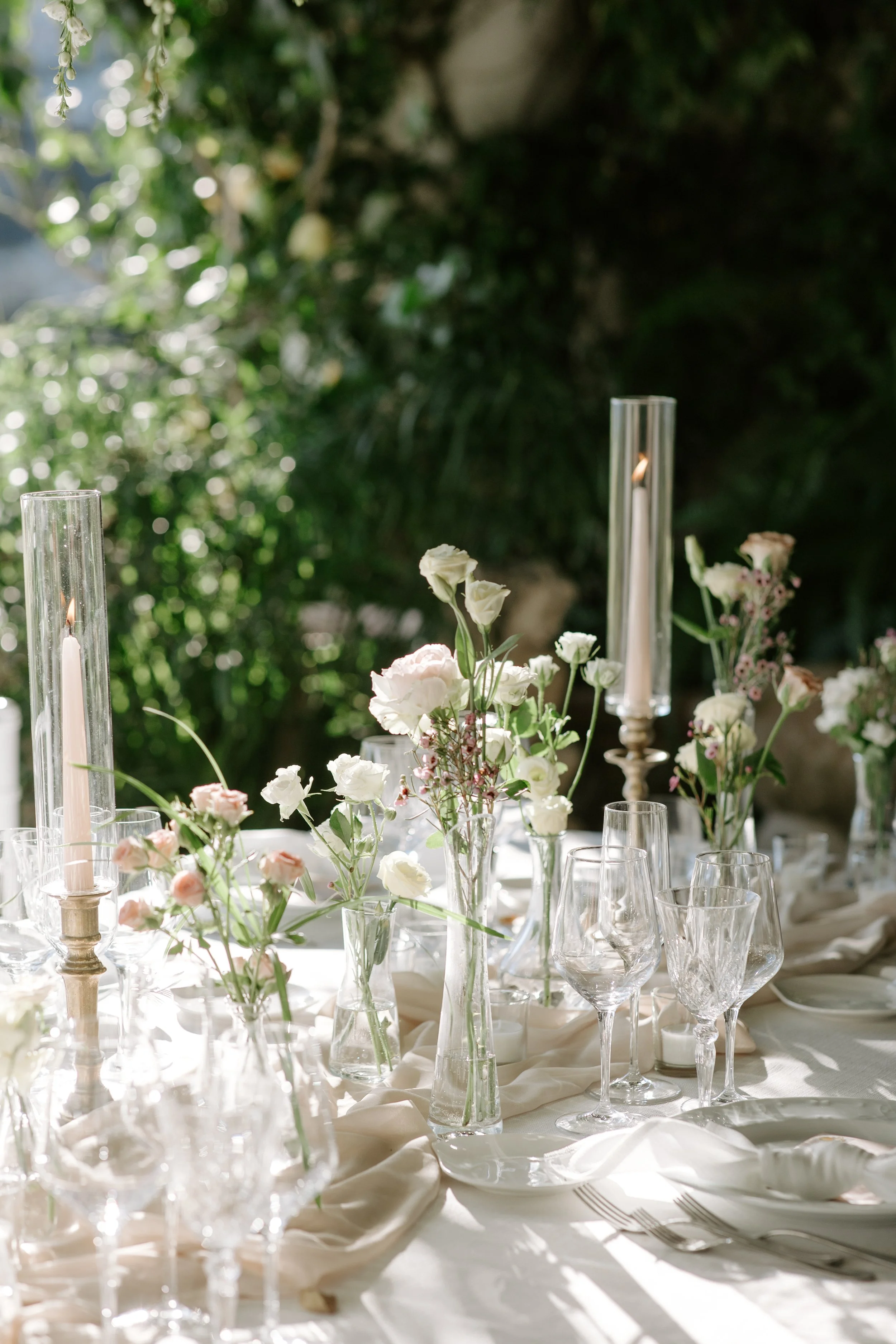 Table setting with clear glass vases filled with white and pink flowers, tall candles in glass holders, and empty wine glasses on a cream-colored tablecloth, outdoors with greenery in the background.