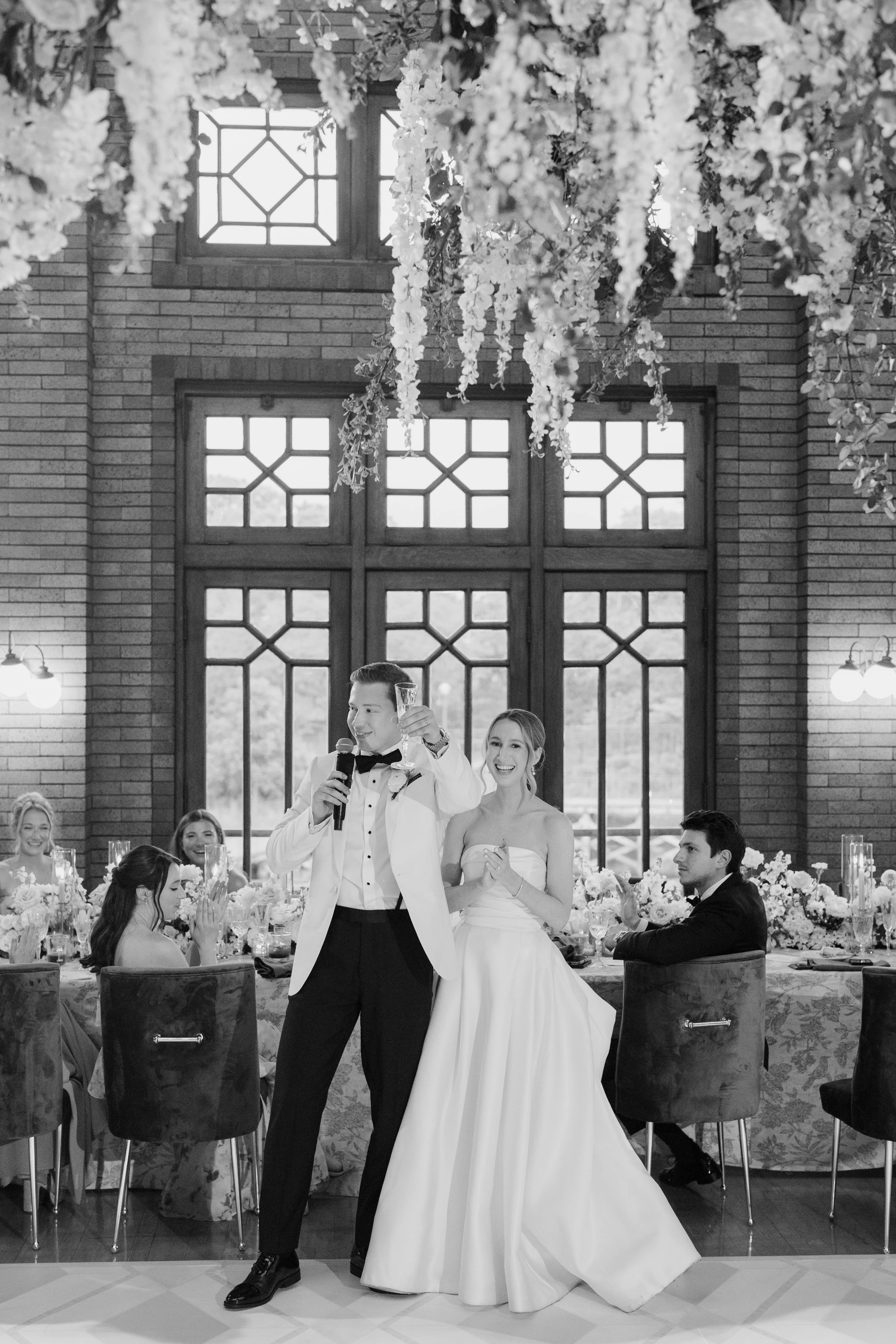 A wedding reception in a grand hall with large windows and brick walls. The groom, wearing a tuxedo, is giving a toast while holding a microphone and a glass. The bride in a strapless wedding gown stands beside him, smiling. Guests are seated at deco