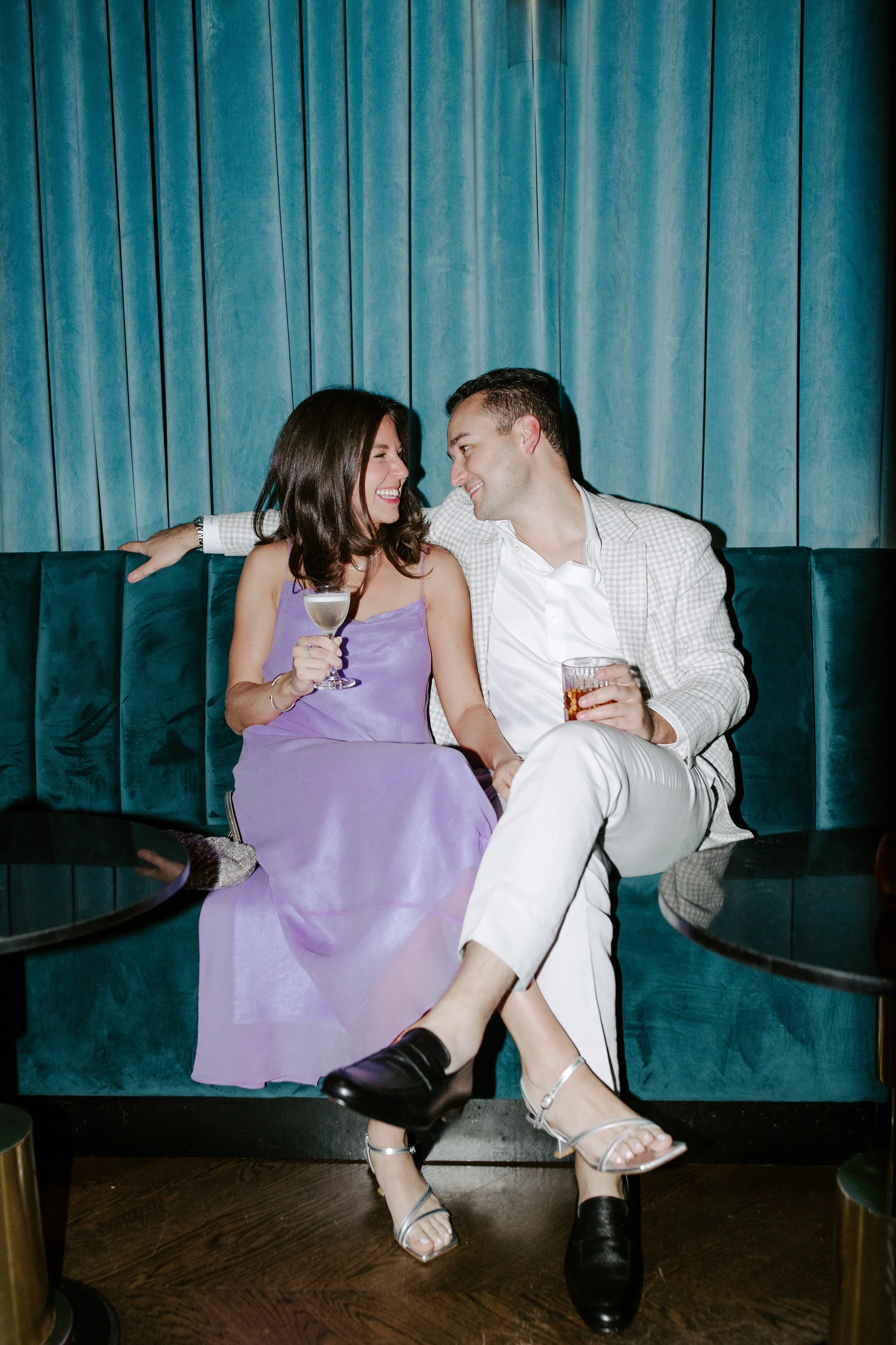 A man and woman sitting close together on a teal velvet sofa, smiling at each other. The woman is holding a cocktail glass, and the man has a glass of soda. They are dressed in formal attire, with the woman wearing a purple dress and the man in a lig