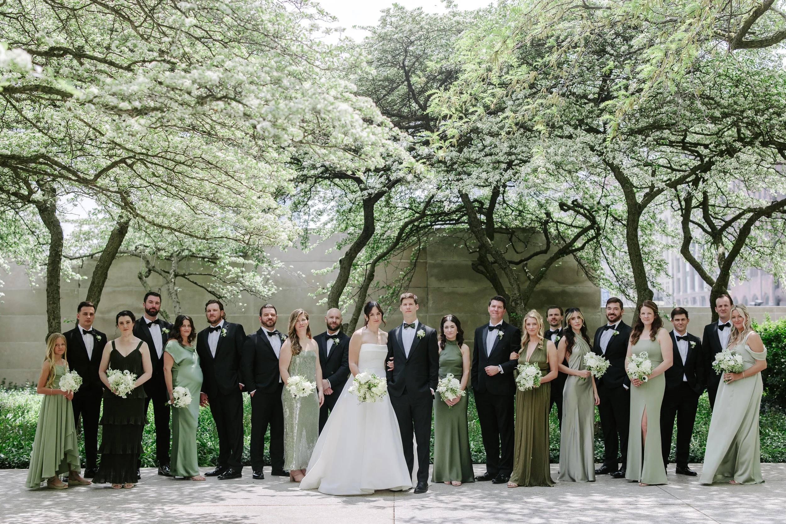A large group of wedding guests and the bride and groom standing outdoors under blooming trees, dressed in formal attire for a wedding ceremony.