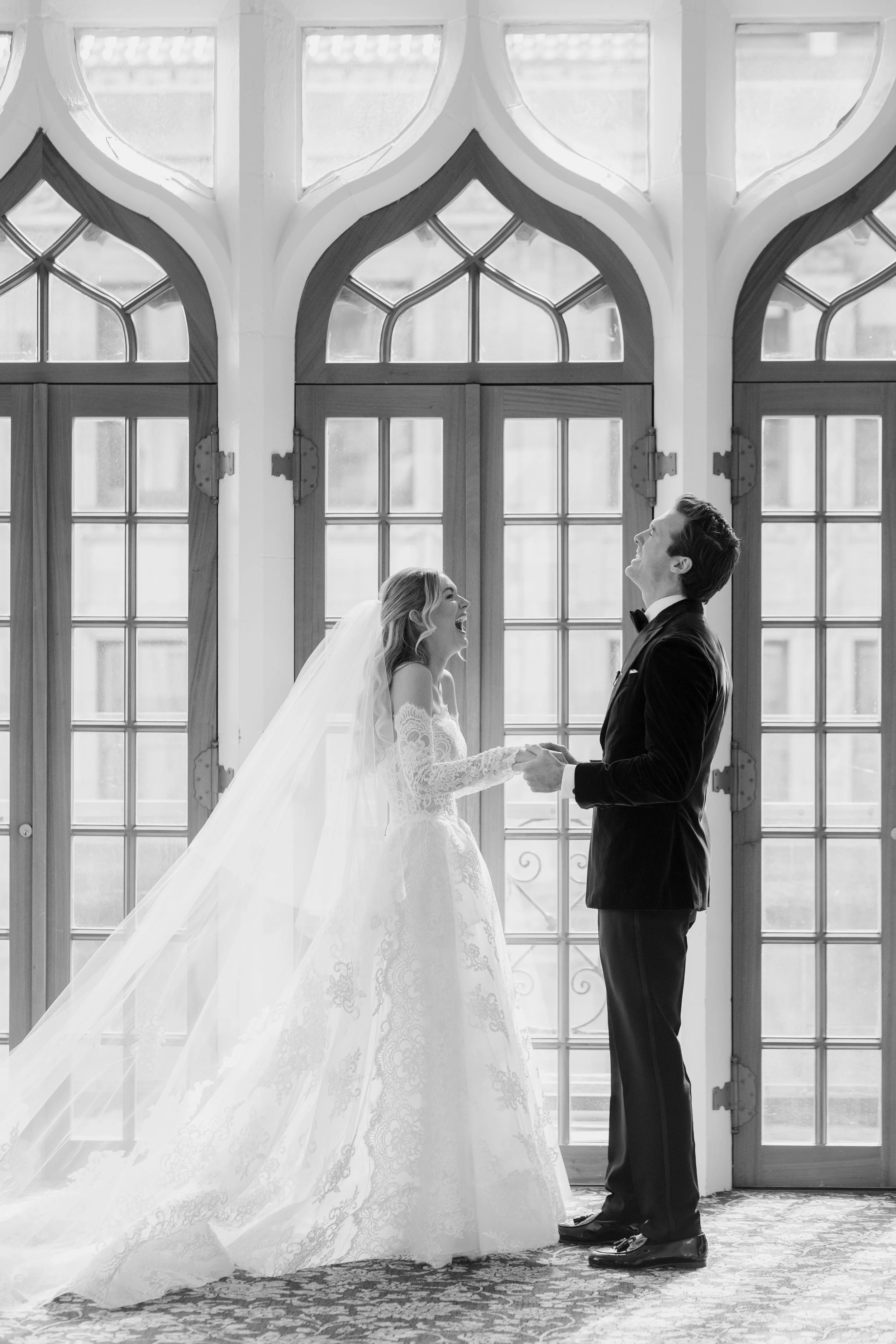 A bride and groom holding hands and smiling at each other inside a room with large gothic-style windows. The bride wears a lace wedding gown and veil, while the groom wears a black tuxedo. The scene is in black and white.