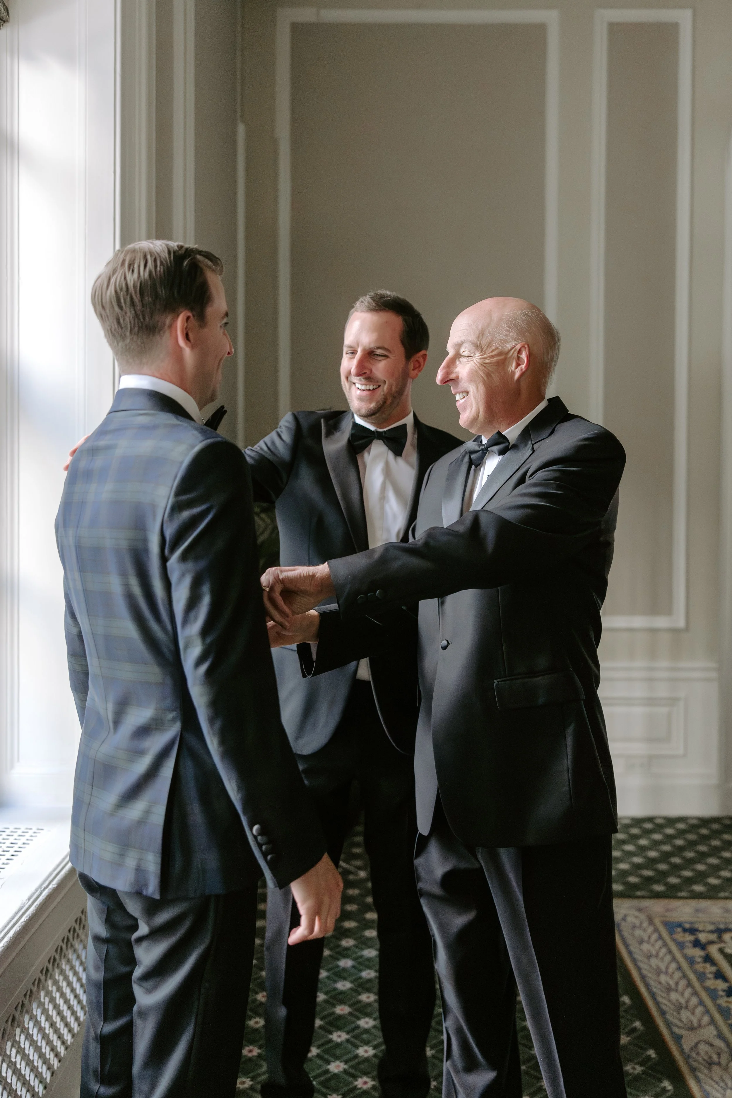 Three men in tuxedos smiling, shaking hands, in a room with patterned carpet and white-paneled walls.