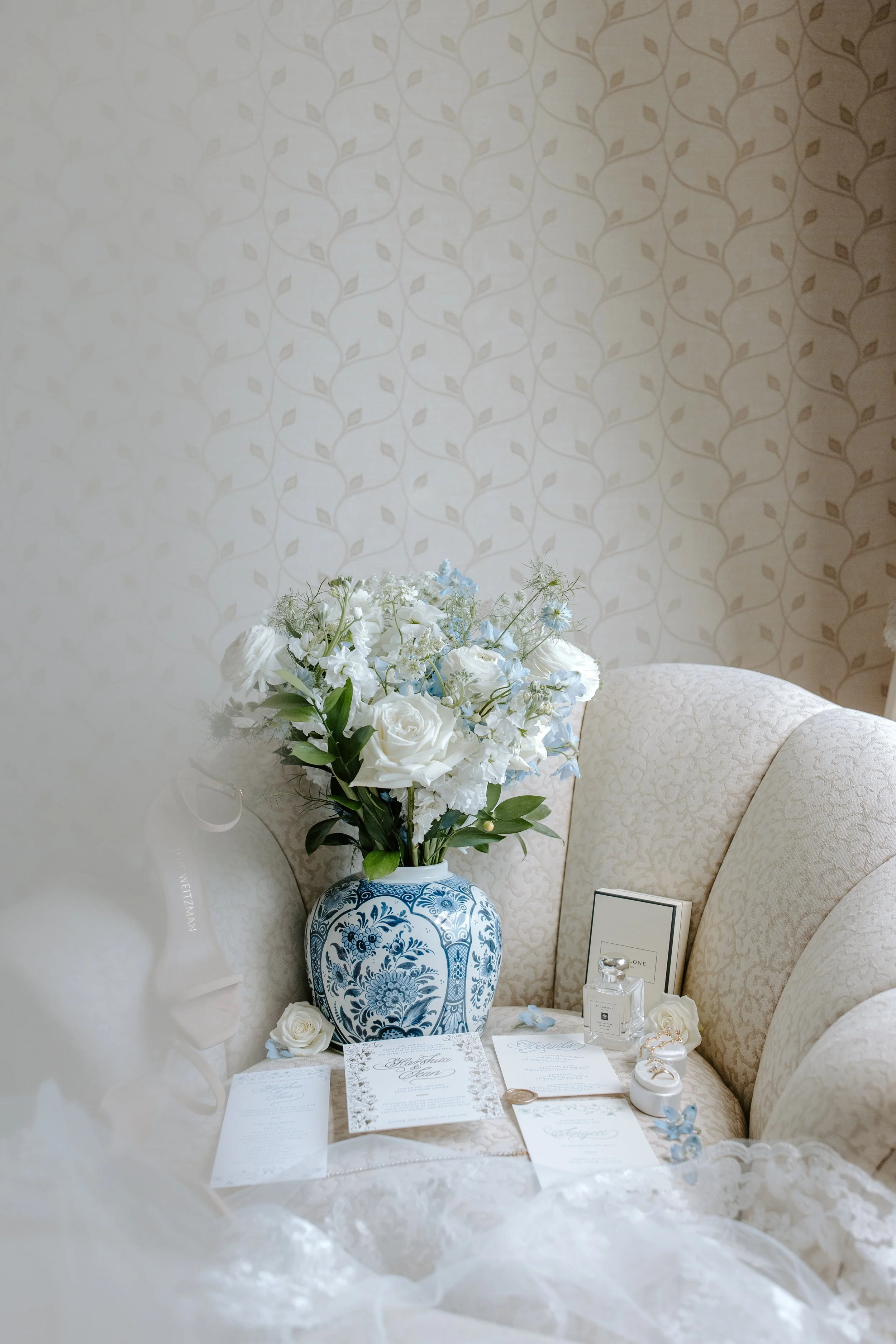 A floral arrangement with white and light blue flowers in a blue and white vase, surrounded by wedding and event stationery, jewelry, and perfume bottles on a cream-colored upholstered armchair with a floral pattern, against a beige wall with a subtl