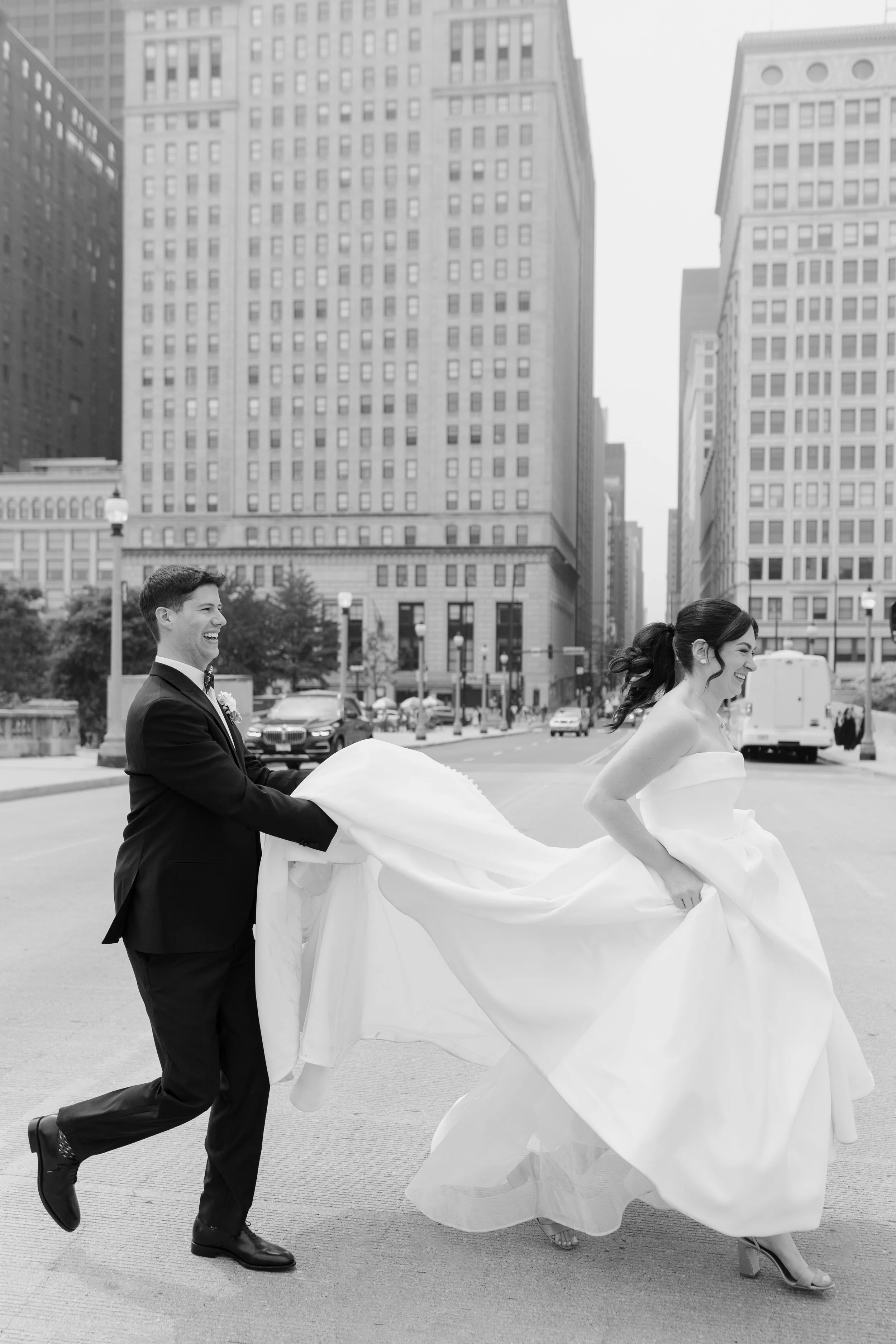 Black and white photo of a groom and bride running on a city street with tall buildings behind them. The groom in a suit is pulling the bride's long wedding dress while she smiles and runs forward.