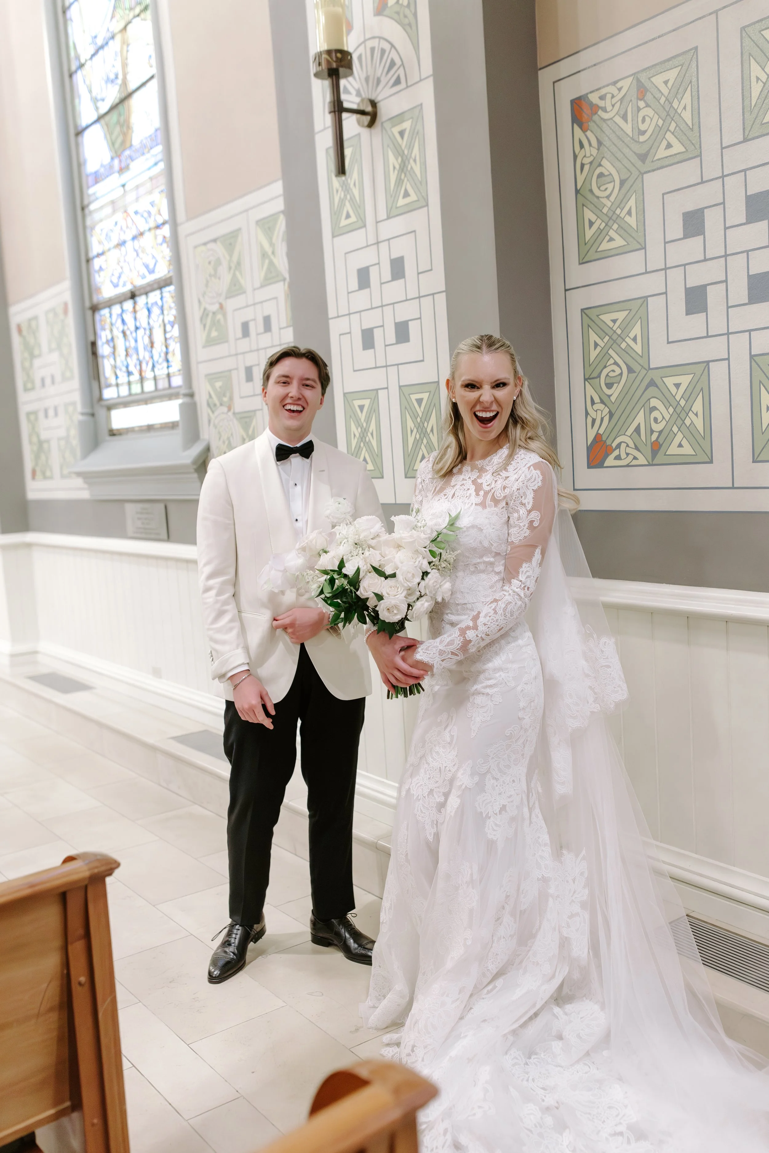 A smiling bride in a lace white wedding dress holding a bouquet of white flowers, standing next to a groom in a white tuxedo with a black bow tie, inside a church with stained glass windows and decorative wall art.