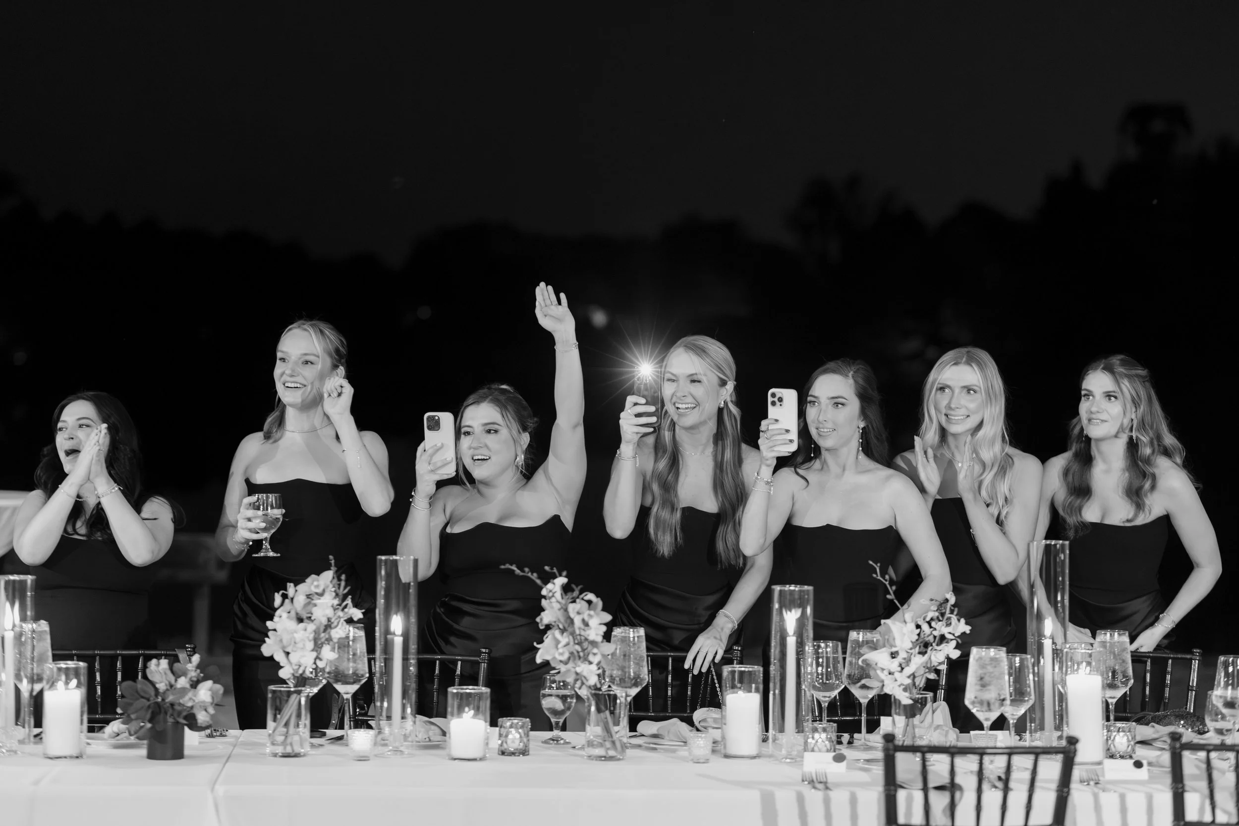 Group of women in formal dresses at a celebration or banquet, raising their hands and holding phones, sitting at a decorated table with candles and flowers, outdoors at night.