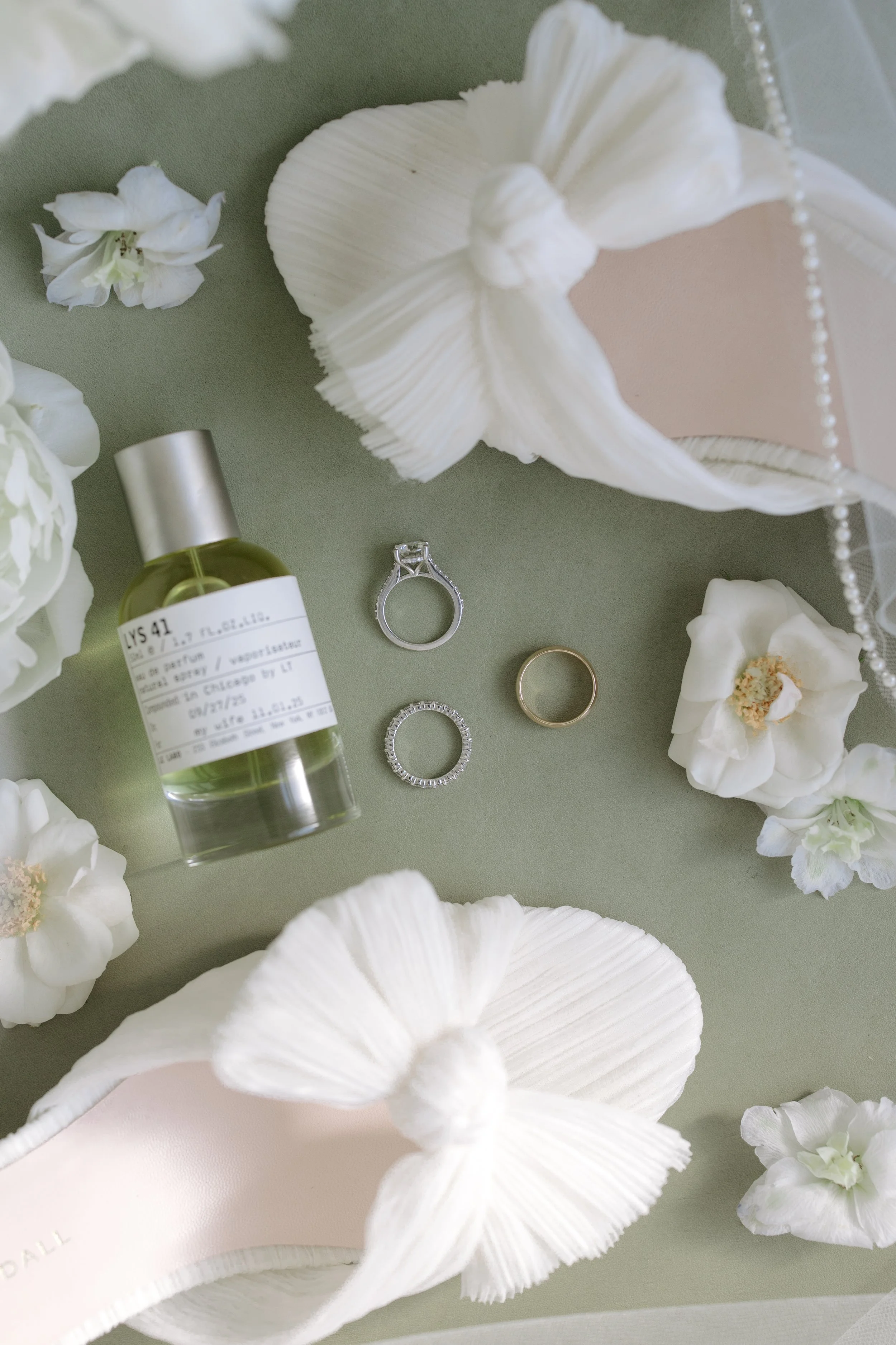 Decorative arrangement featuring white flowers, a ring, a perfume bottle, and jewelry on a green surface.