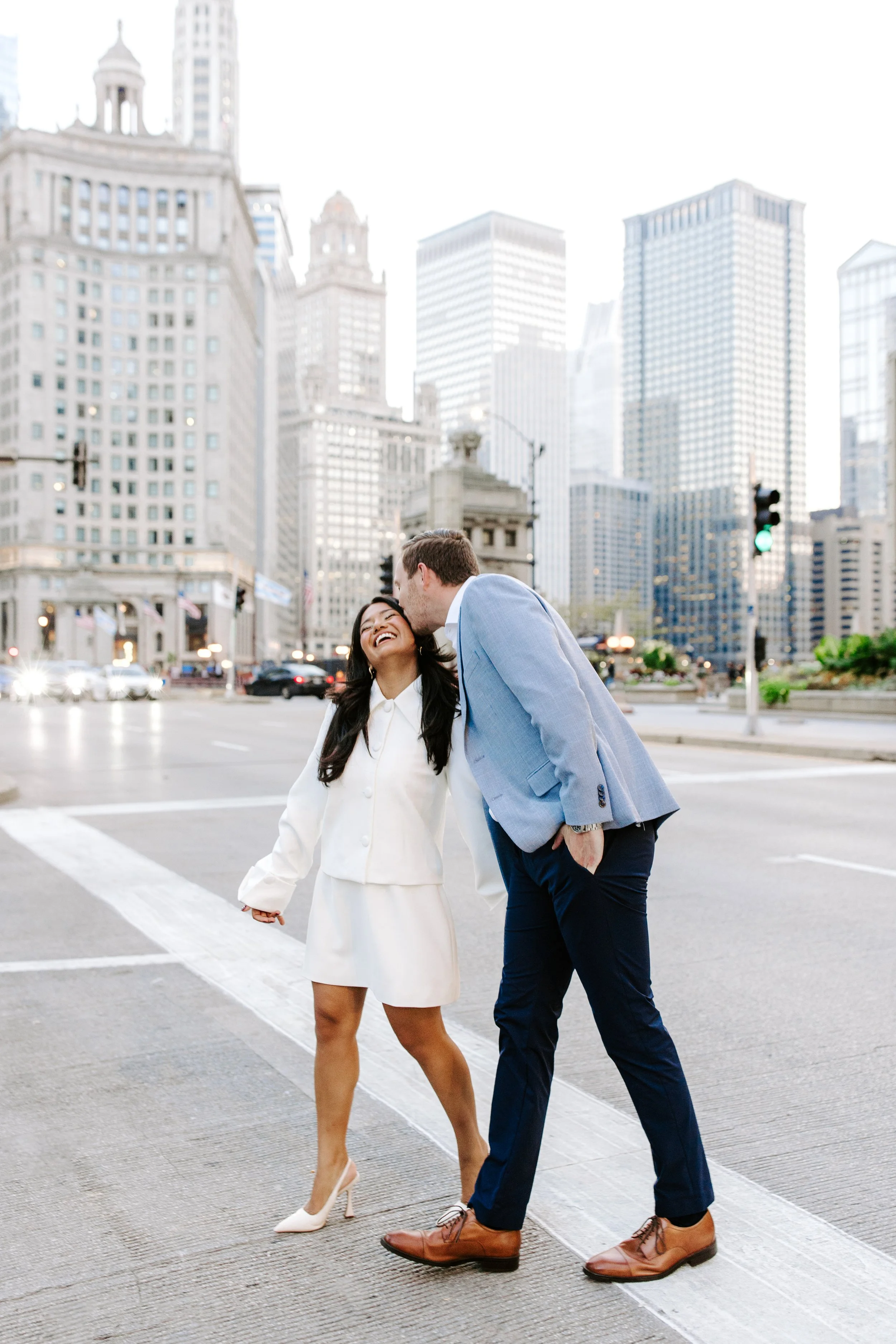 A couple holding hands and walking across a busy city crosswalk, with tall skyscrapers in the background. The man is leaning to kiss the woman's temple.