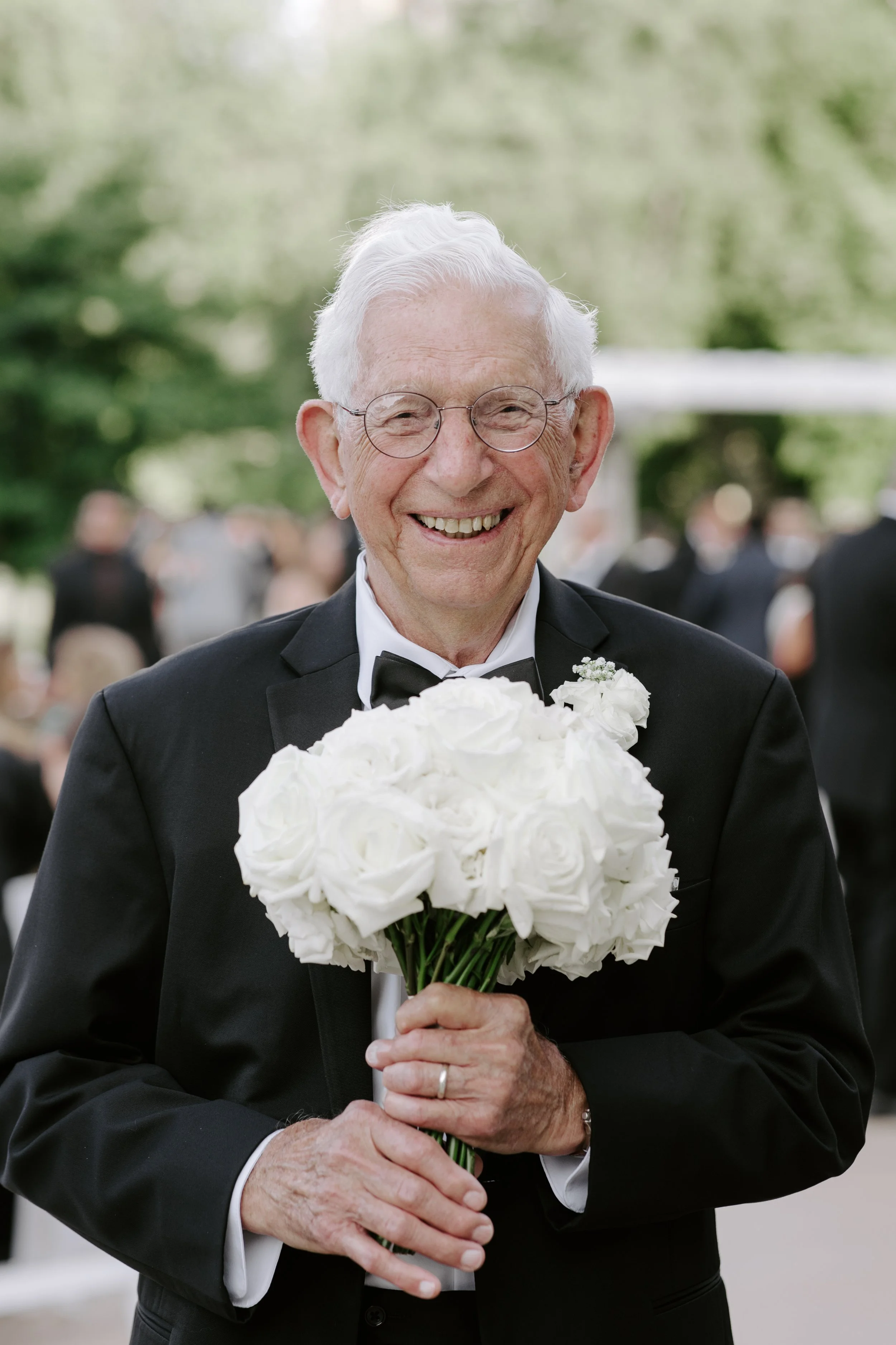 Older man in a tuxedo holding a bouquet of white roses, smiling at an outdoor event with blurred people and trees in the background.