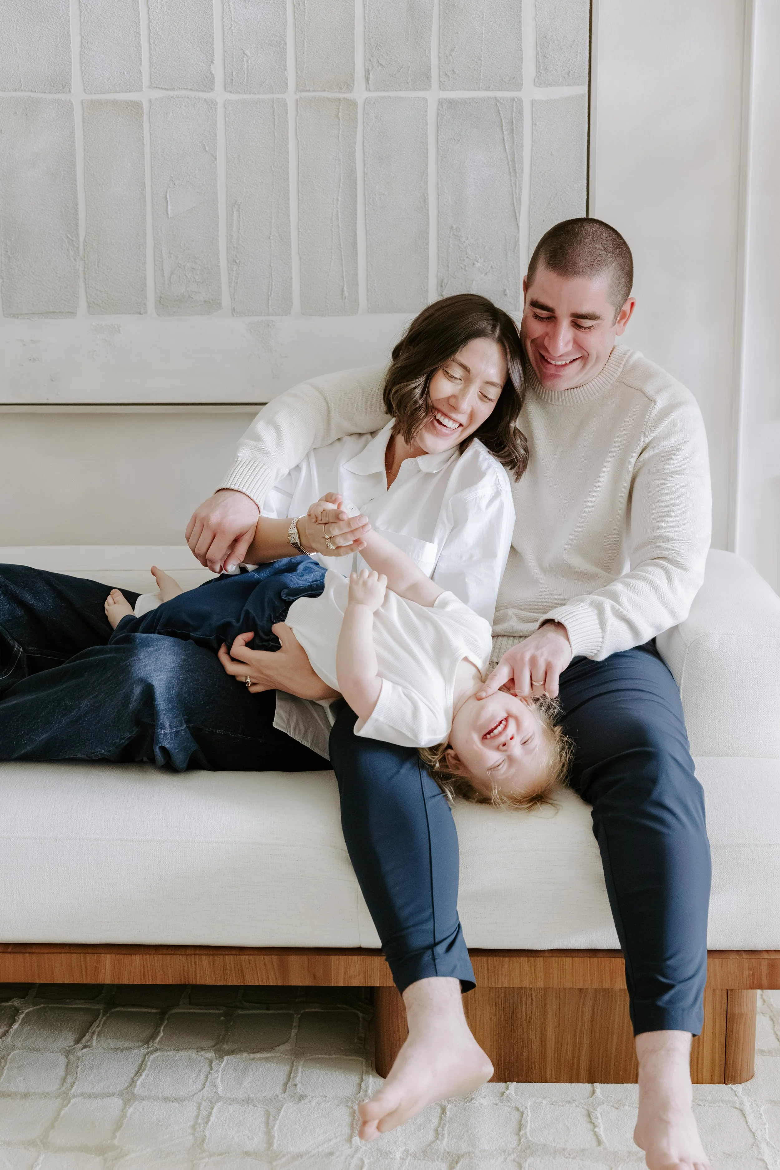 A happy family with a woman, a man, and two children sitting on a sofa, smiling and playing together in a cozy home setting.