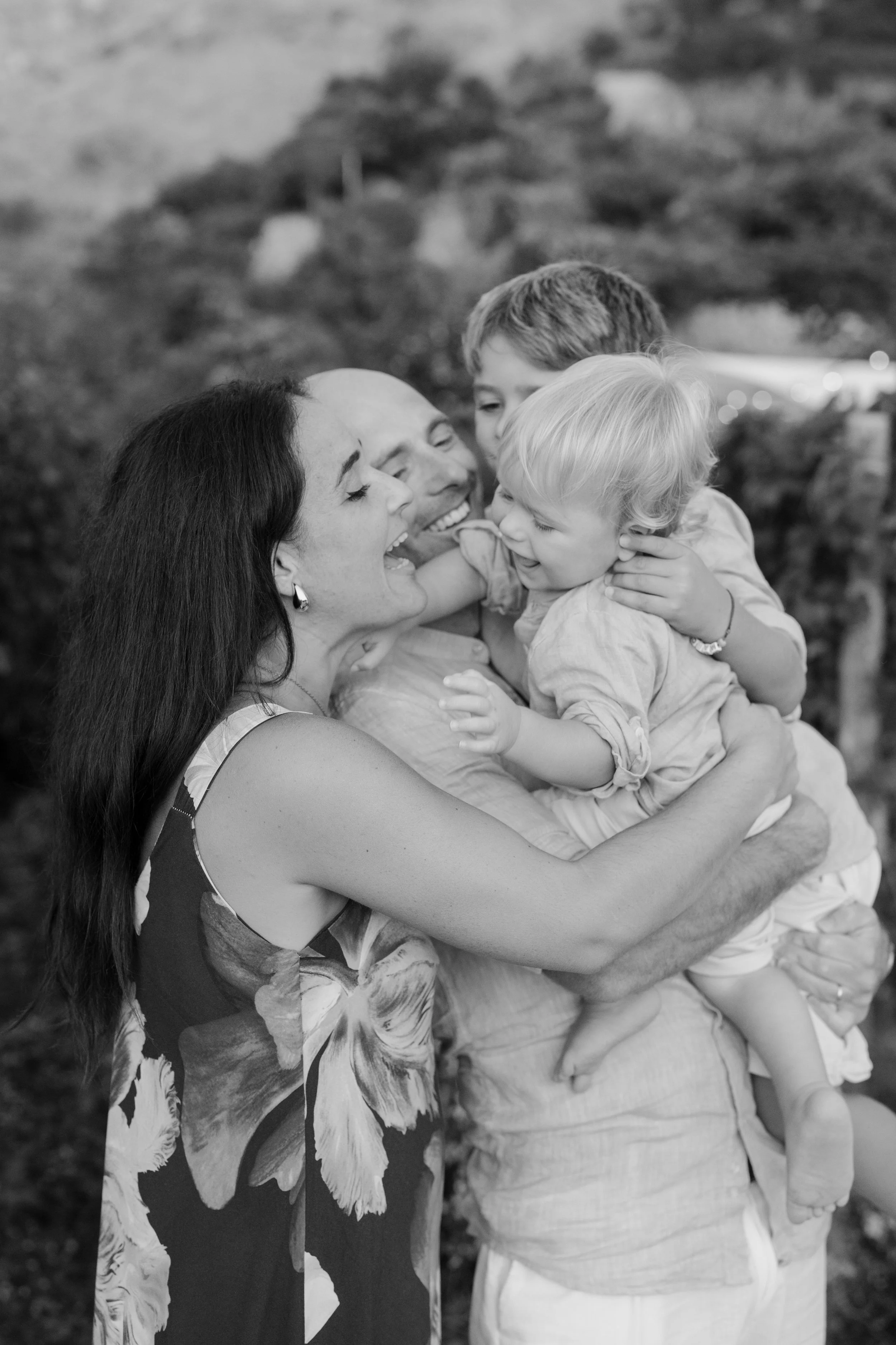A black and white photo of four people, including two women and two children, hugging and laughing outdoors.