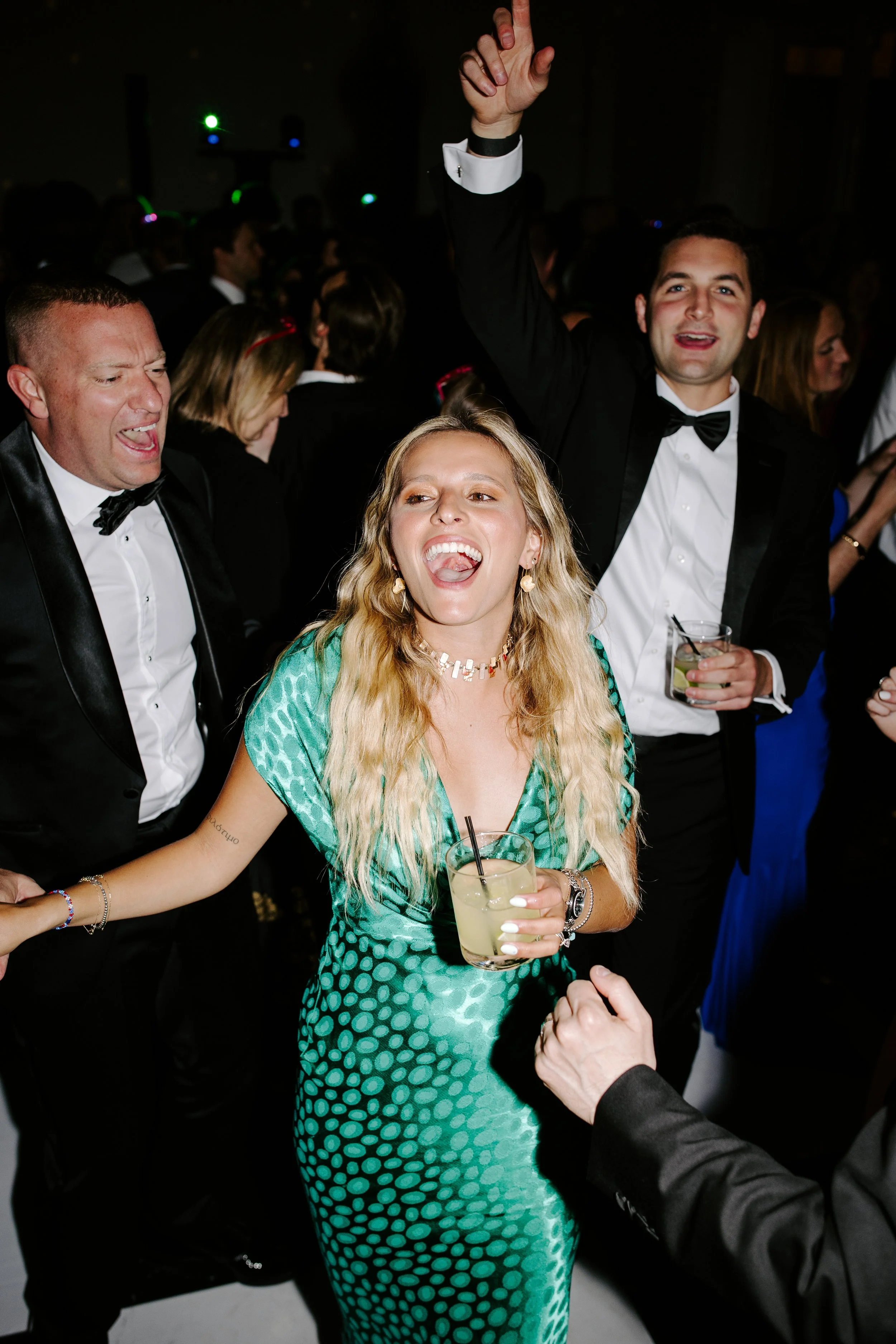 A group of people dressed in formal attire celebrating at a party or event. A woman in a green dress with a polka dot pattern holds a drink and appears to be having fun, while a man in a tuxedo with a bowtie is holding a drink and smiling. Other gues