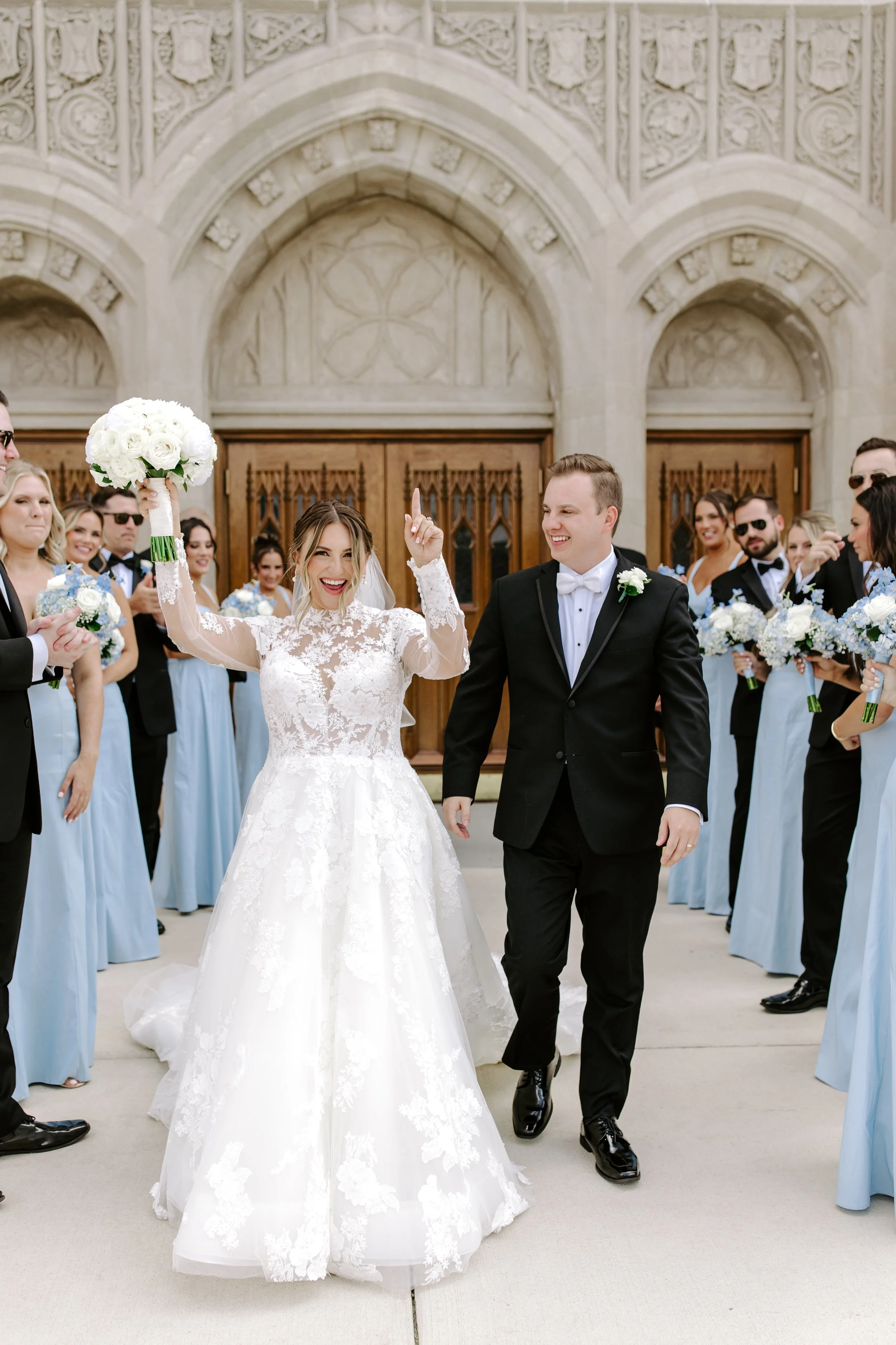 Bride celebrating with her wedding bouquet raised, surrounded by wedding party and guests outside church building.