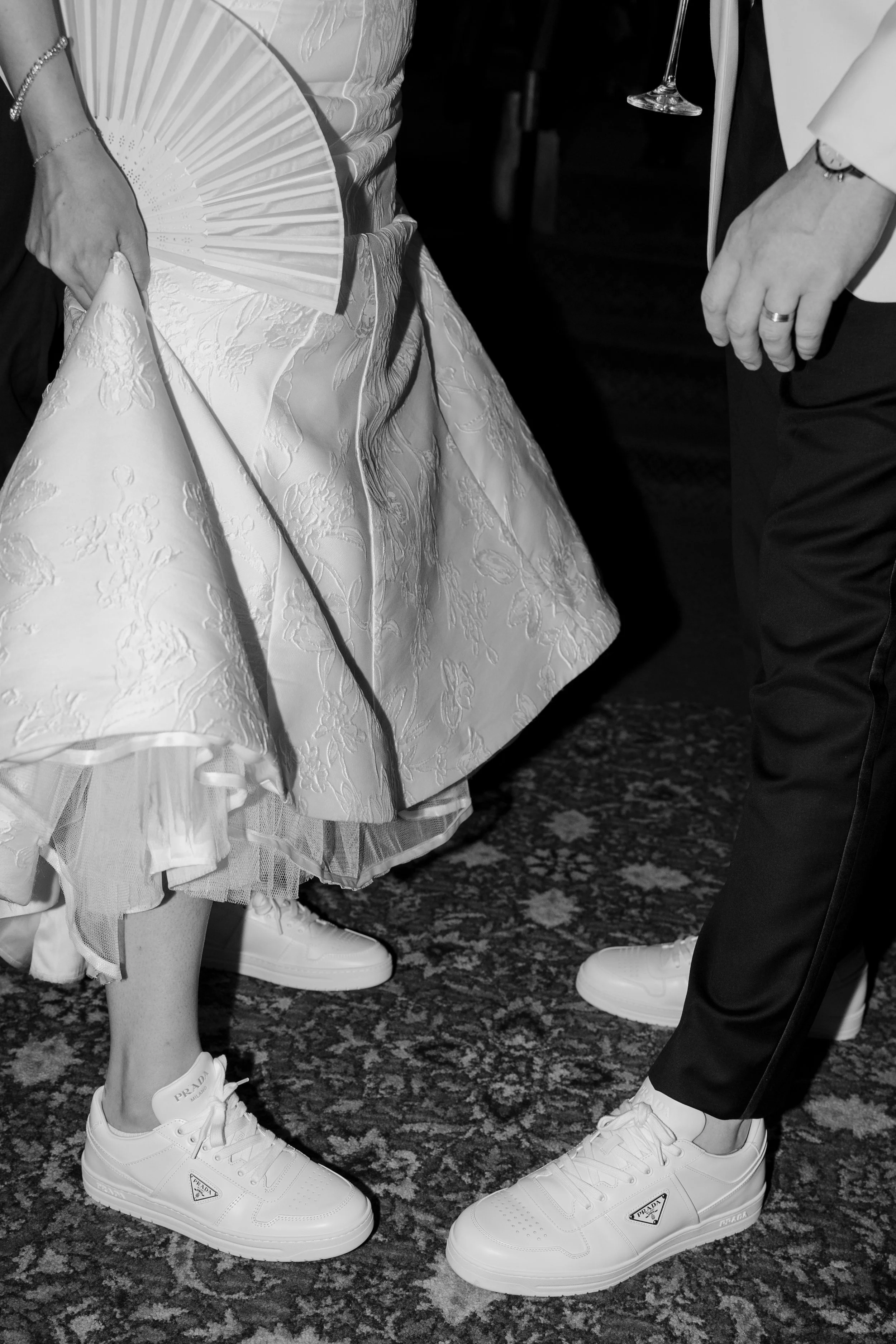 Close-up of a couple's feet and lower bodies at a formal event, with the woman wearing a floral dress with a tulle hem, white sneakers with Prada branding, and holding a fan; the man wearing dark dress pants, a white blazer, and white sneakers. They 