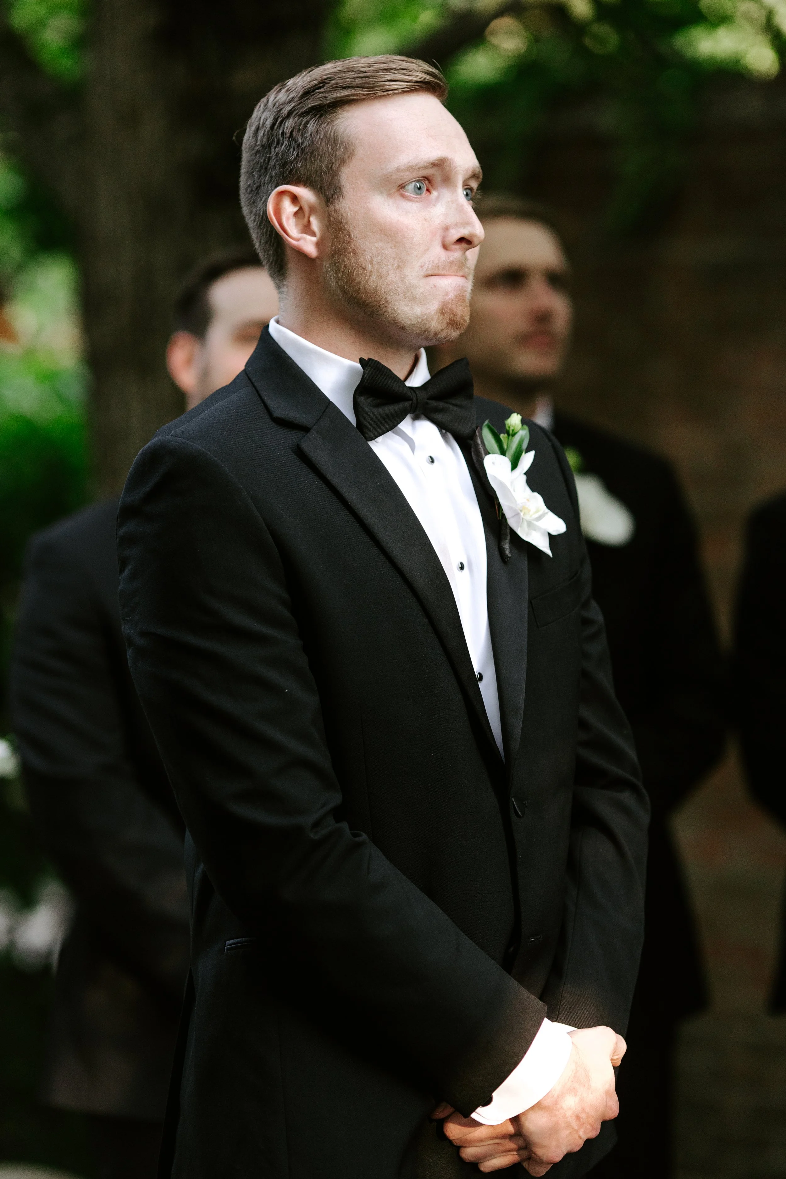 Man wearing a tuxedo with a bowtie and a white flower boutonniere standing with hands clasped, during a formal event or wedding ceremony outdoors.