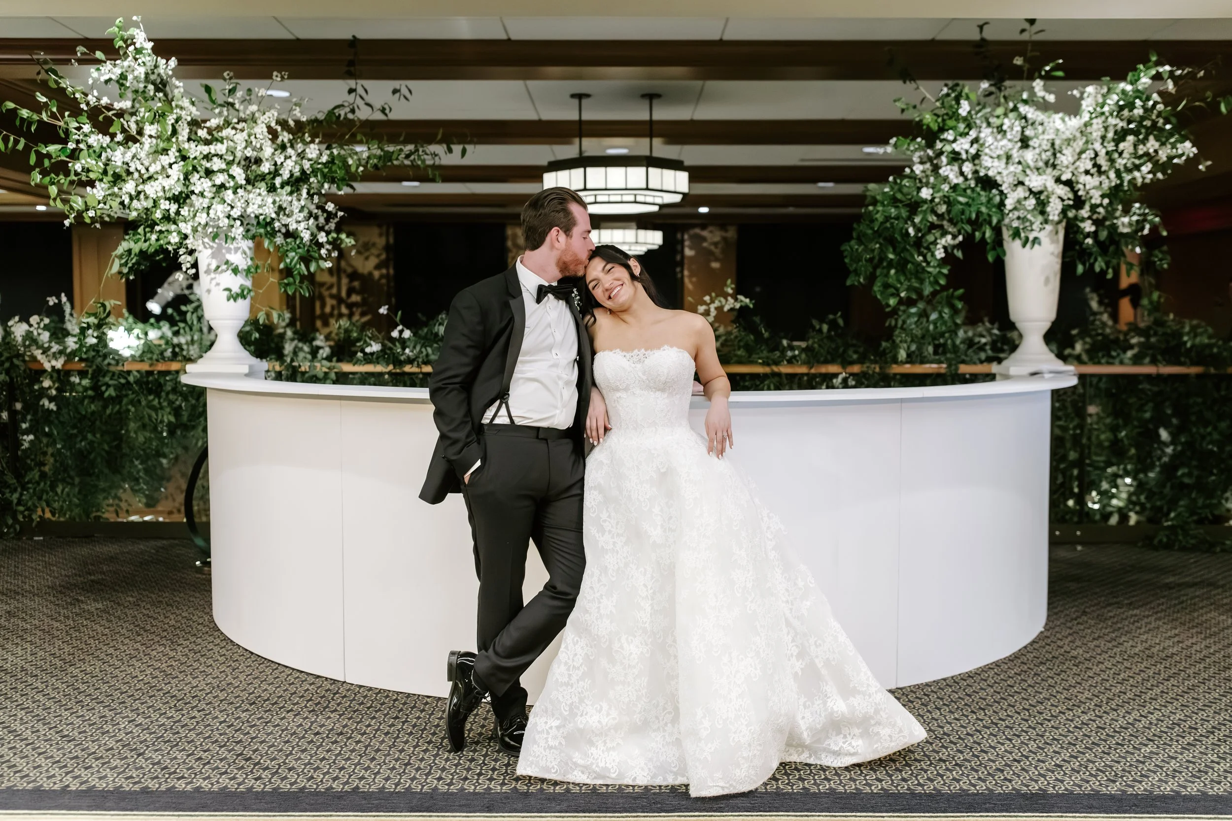 A couple dressed in wedding attire standing in front of large floral arrangements with smiling and close affectionate pose.