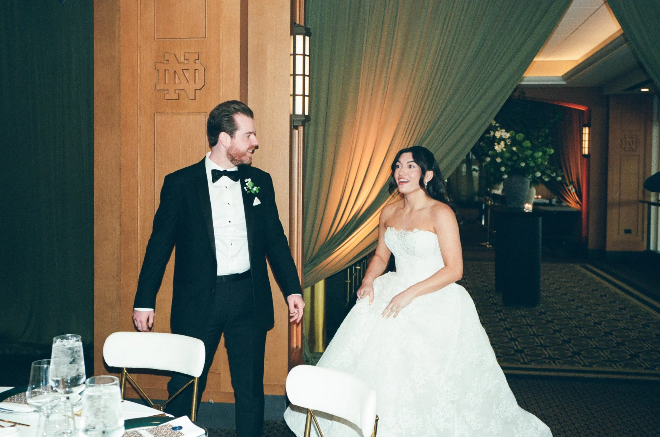 A bride and groom holding hands during their wedding reception in an elegant indoor venue with decorated walls and floral arrangements.