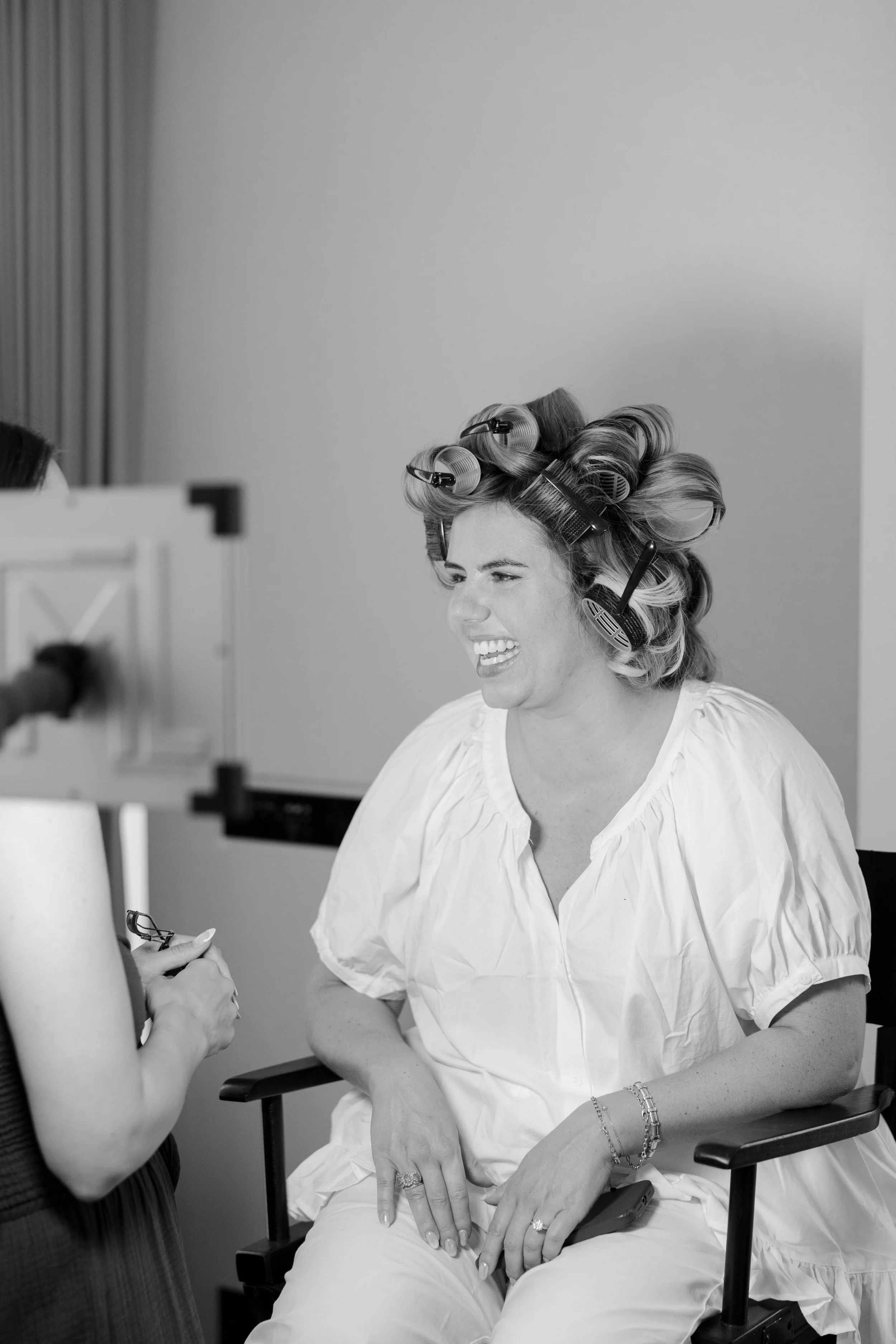 A woman with curlers in her hair, sitting in a chair, smiling and looking at someone outside the frame, while another person adjusts her clothing.