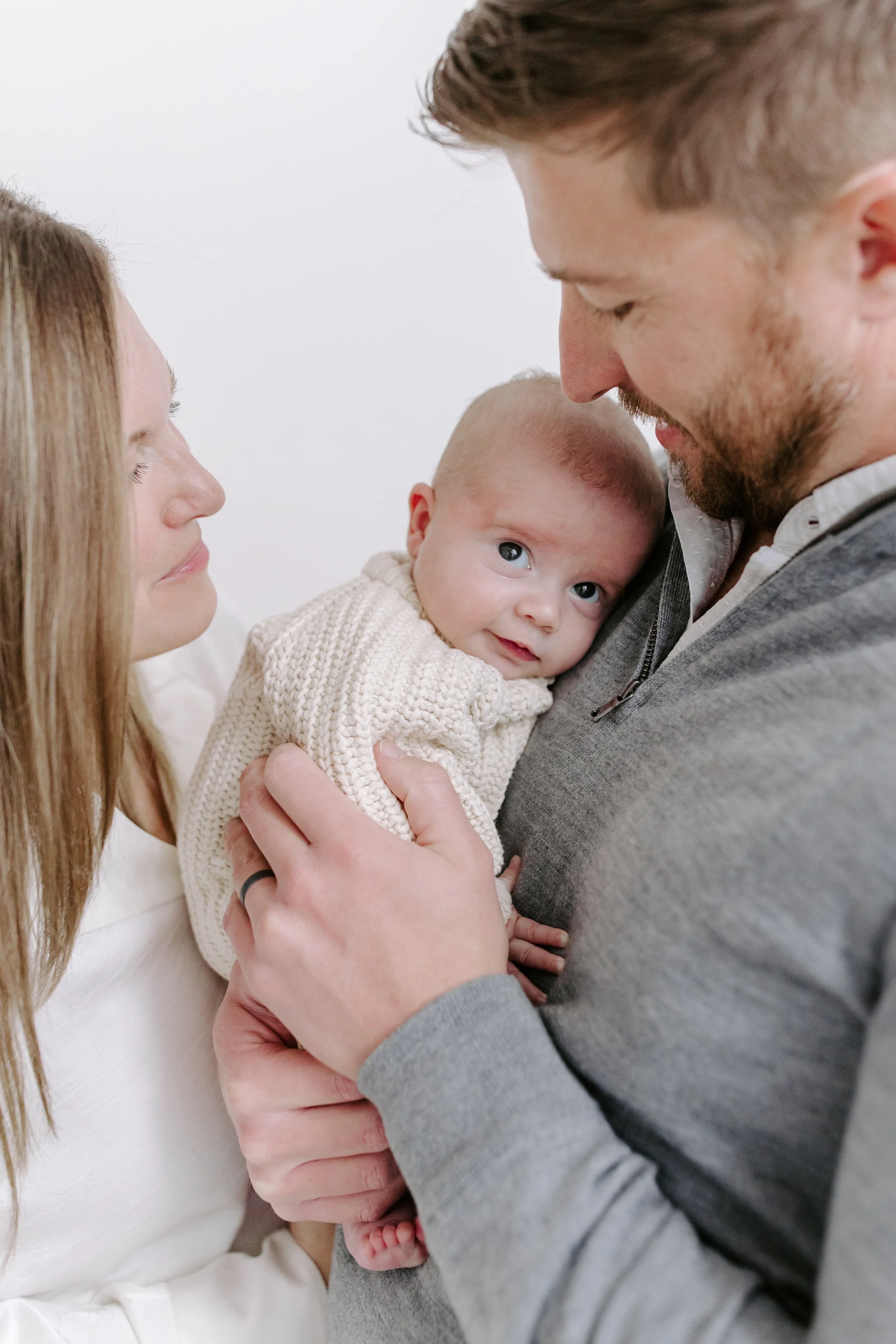 A family of three cherishing a moment together with a young baby in the father's arms, as the mother looks lovingly at them.
