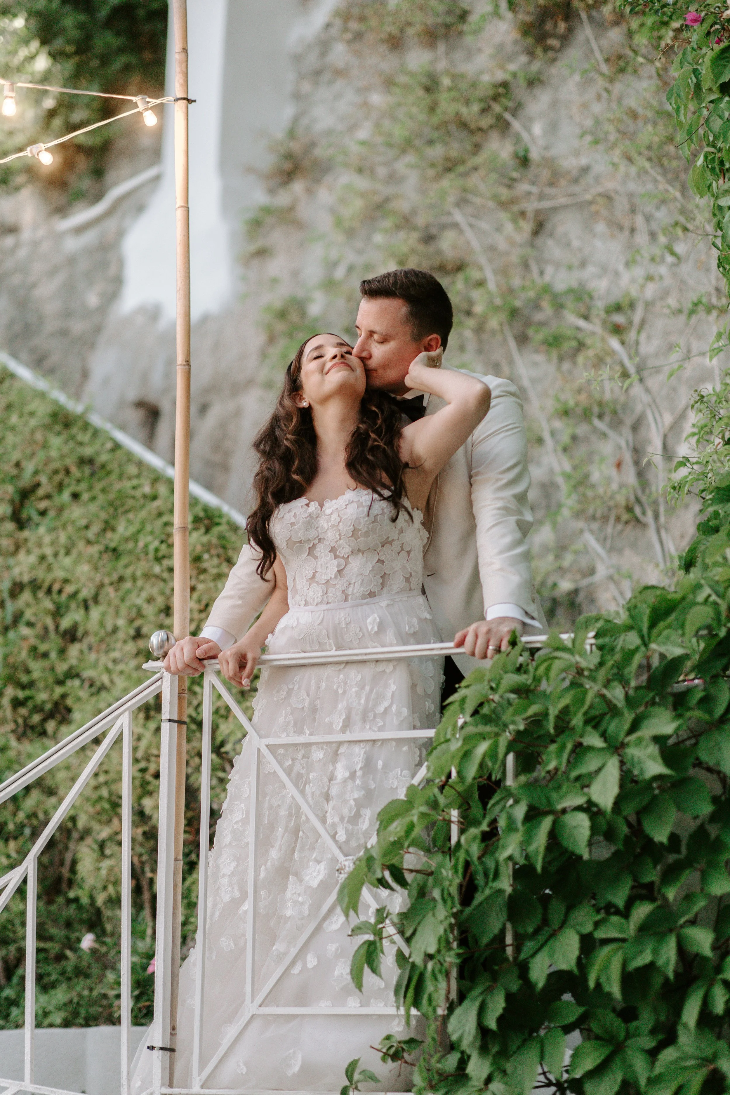 A bride and groom on a balcony decorated with string lights, surrounded by greenery and a rocky hillside.
