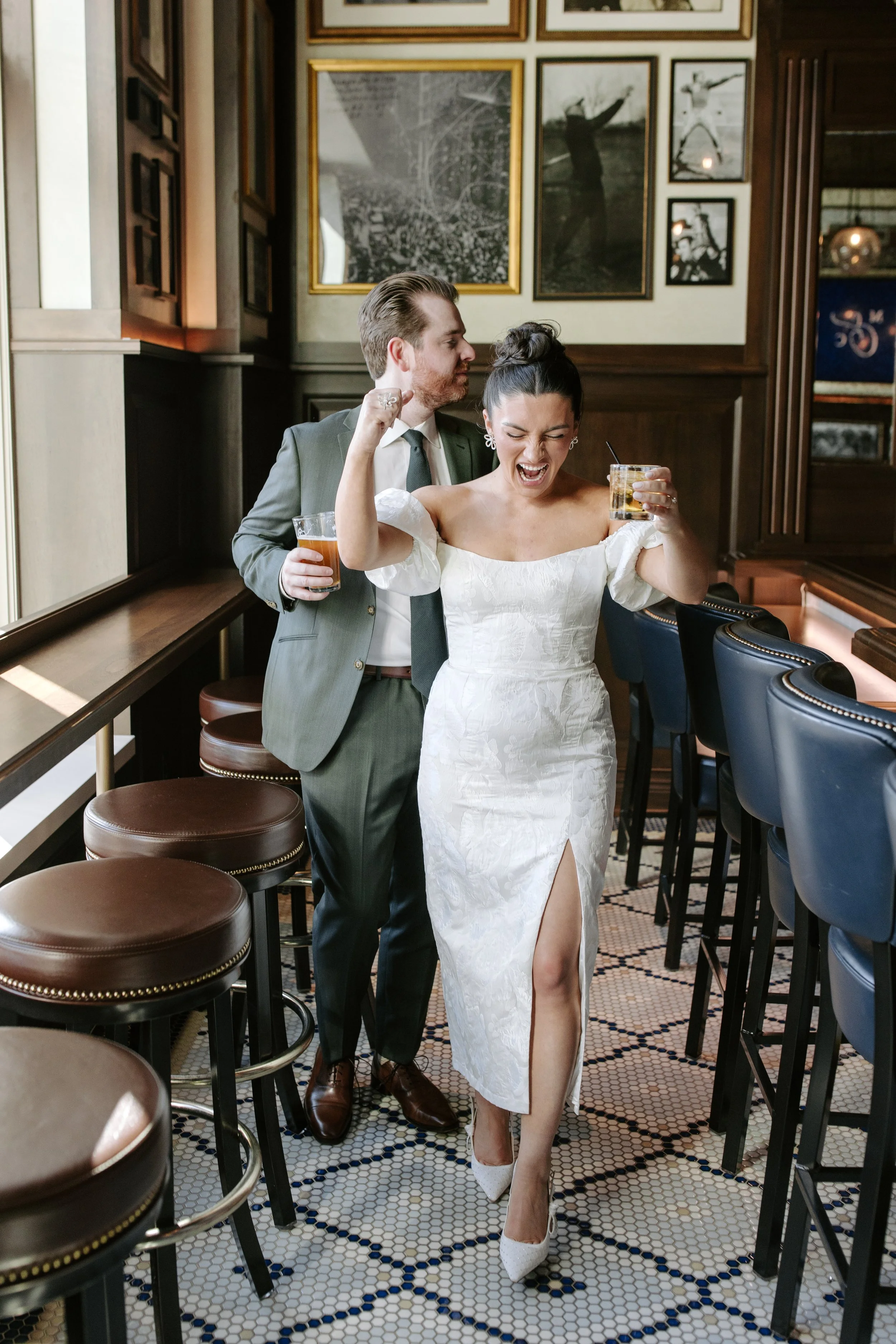 A woman in a white dress with a high slit dancing in a bar or restaurant, holding a drink in one hand and raising her fist in celebration, while a man in a suit stands behind her also holding a drink, with framed photos on the wall behind them.