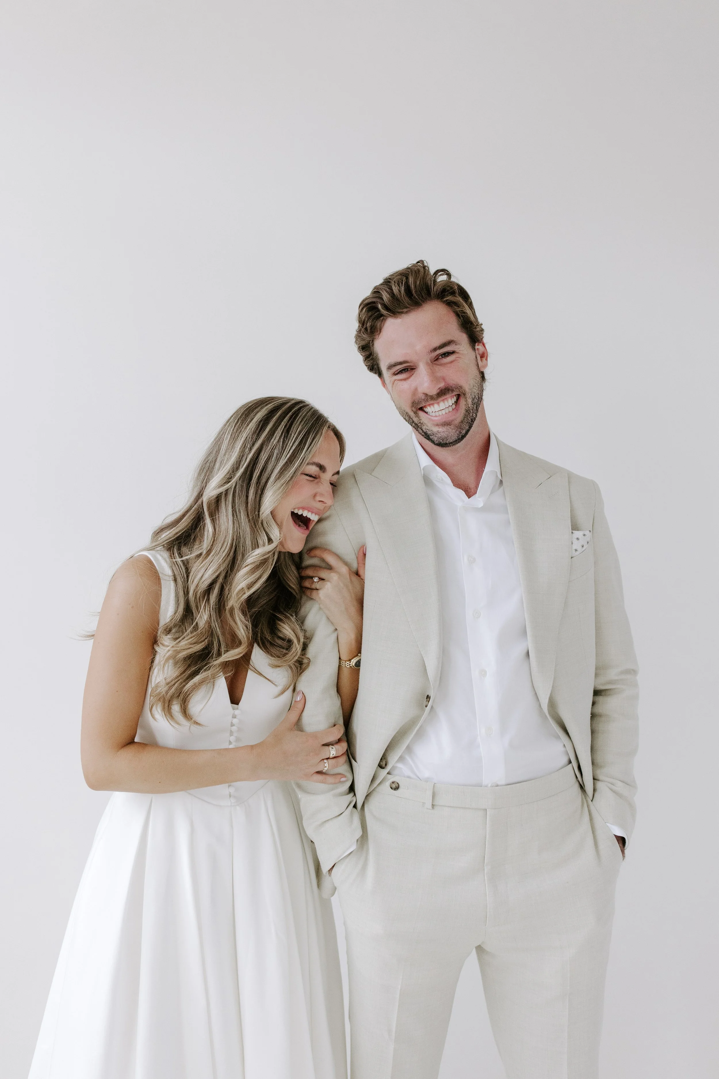 A smiling man in a beige suit with a white shirt stands next to a woman laughing, wearing a white dress, against a plain white background.