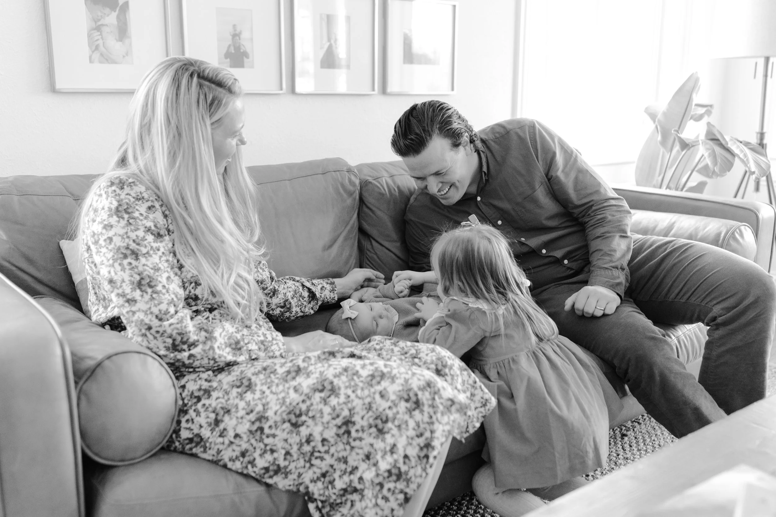 A black and white photo of a family gathered on a couch in a living room. Two young children are looking at a baby lying on a pillow with a breathing tube while the parents are smiling and holding the children's hands.