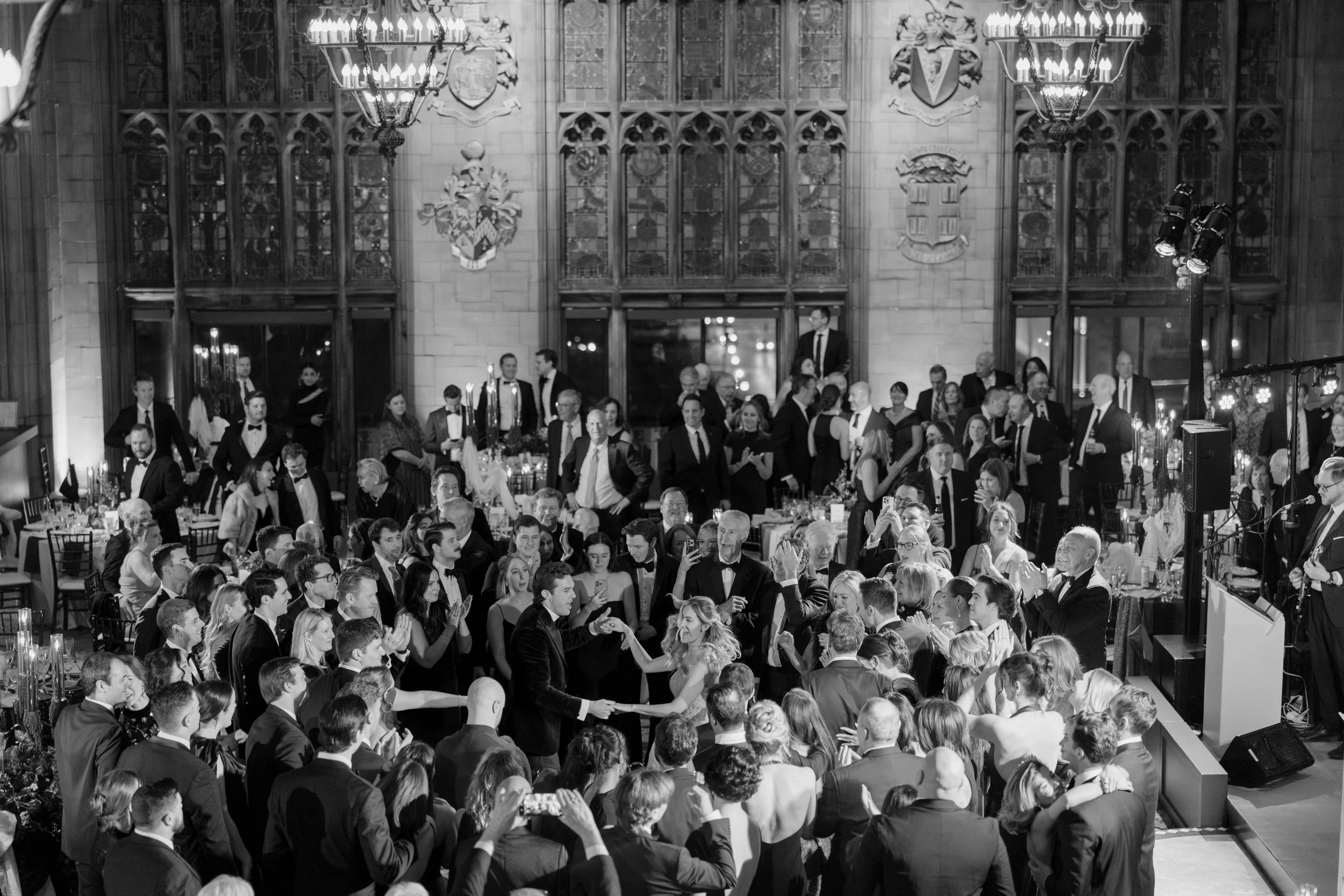 A black and white photo of an elegant banquet hall filled with well-dressed people, with chandeliers hanging from the ceiling, and coats of arms displayed on the wall.