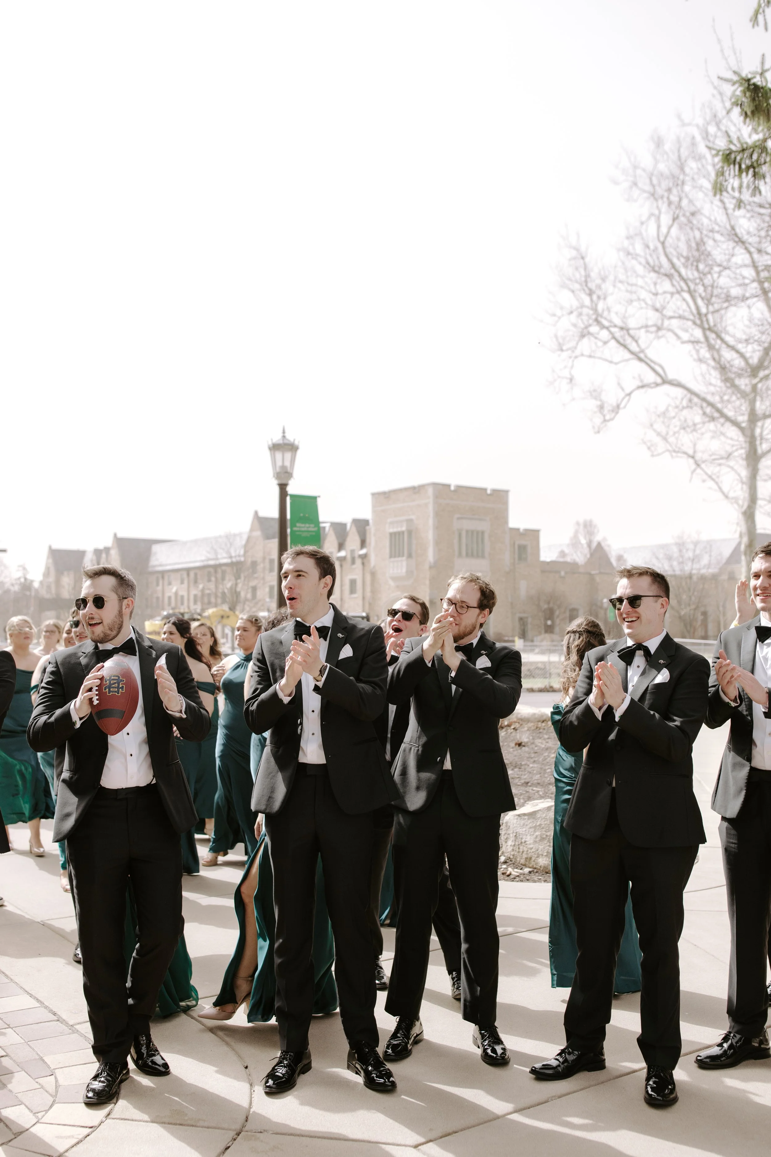 A group of men in tuxedos, some holding drinks and wearing sunglasses, gathered outdoors during daytime with buildings and trees in the background.