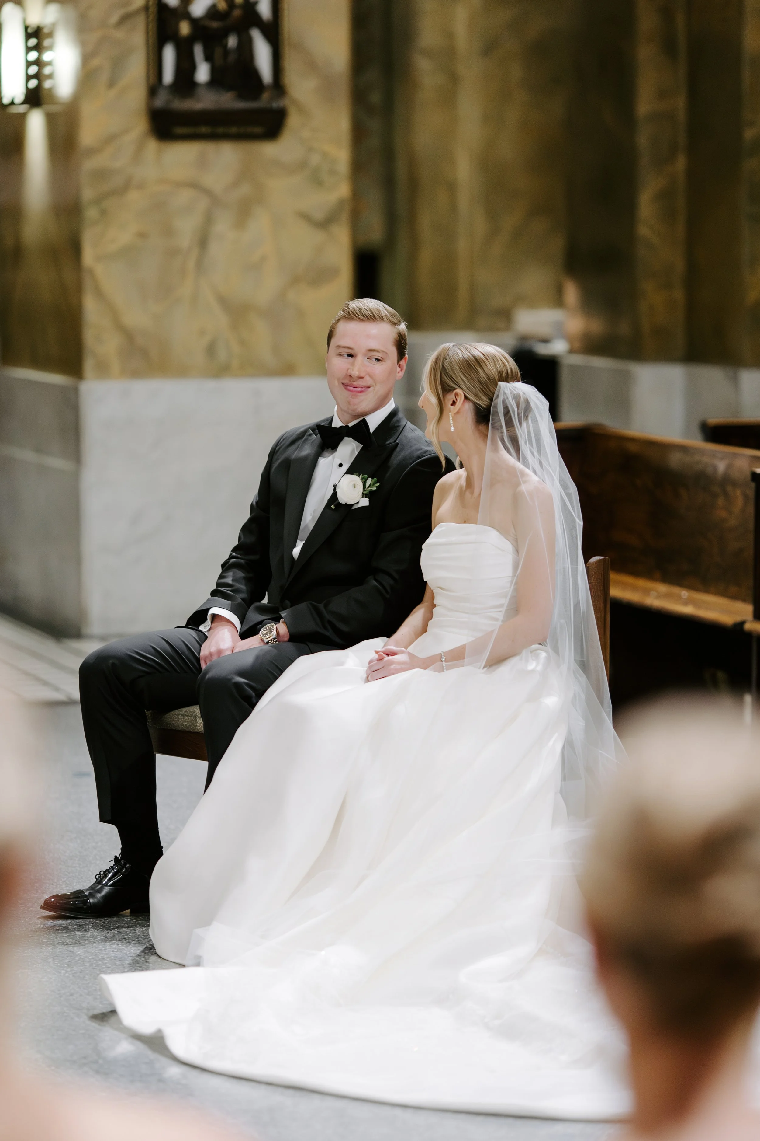 A bride and groom sitting in a church, looking at each other with smiles. The groom is wearing a black tuxedo with a bow tie and a white boutonniere, while the bride is in a strapless white wedding gown with a veil. The background features stone wall