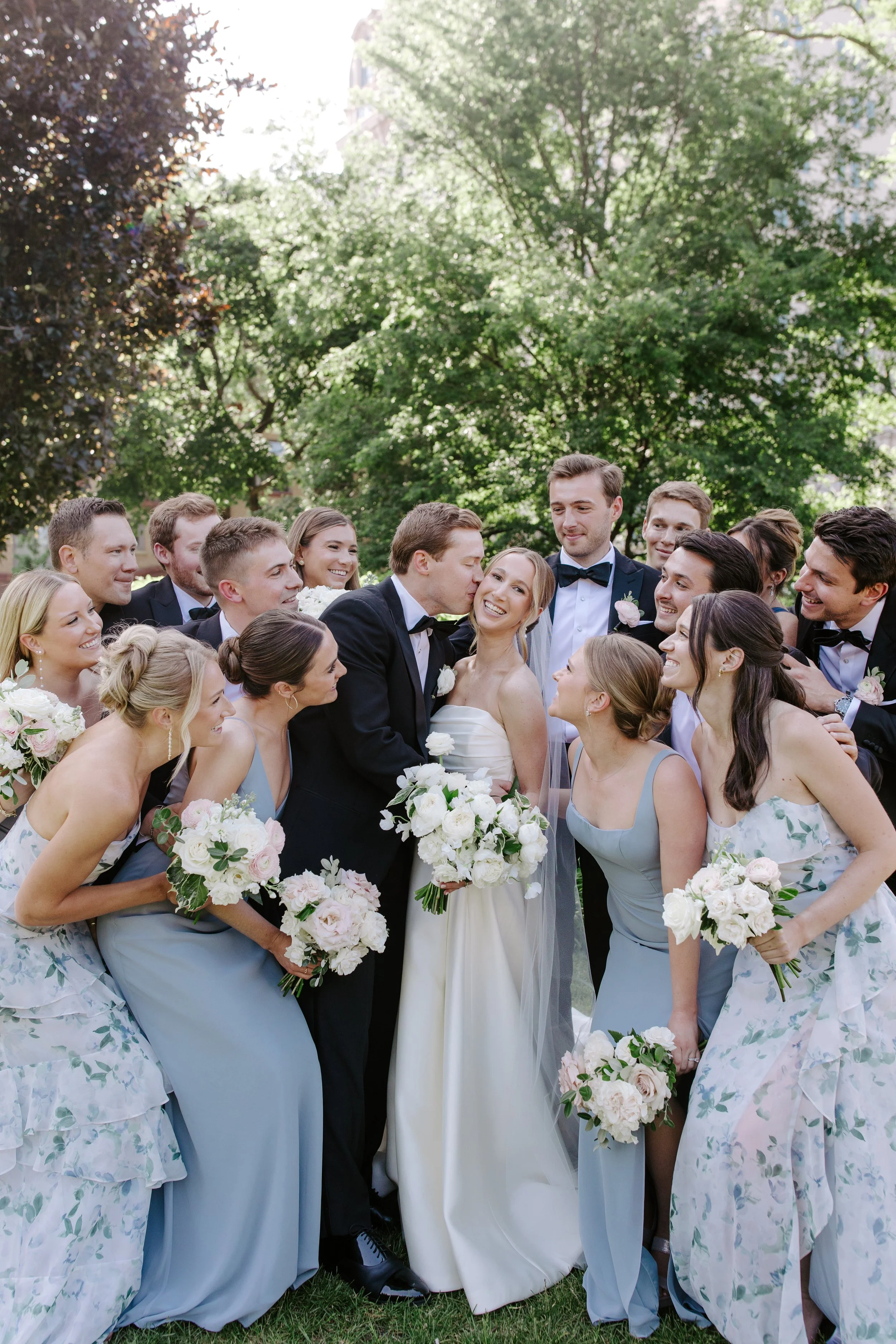 A group of people dressed in wedding attire gathered outdoors, with the bride and groom at the center. The bride is wearing a white wedding dress, and the groom is in a black tuxedo. Both are smiling, surrounded by bridesmaids in blue dresses holding