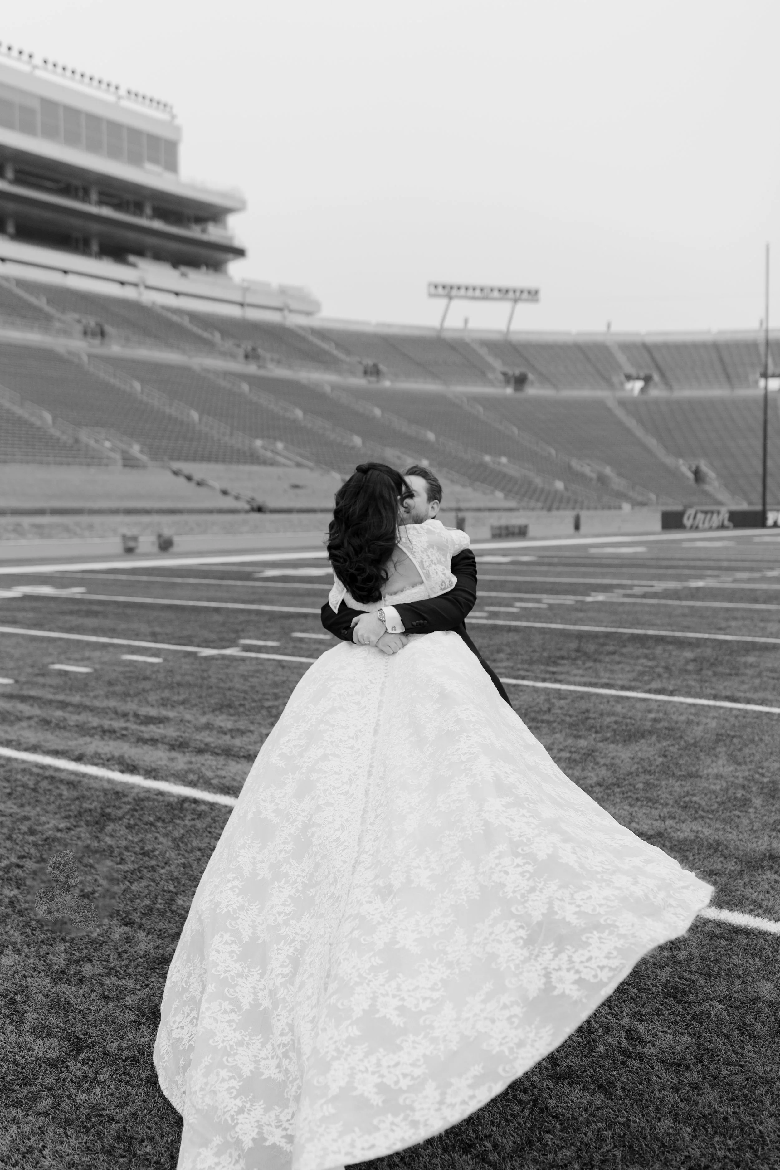 A bride and groom standing on a football field, embracing each other, with empty stadium seats in the background.