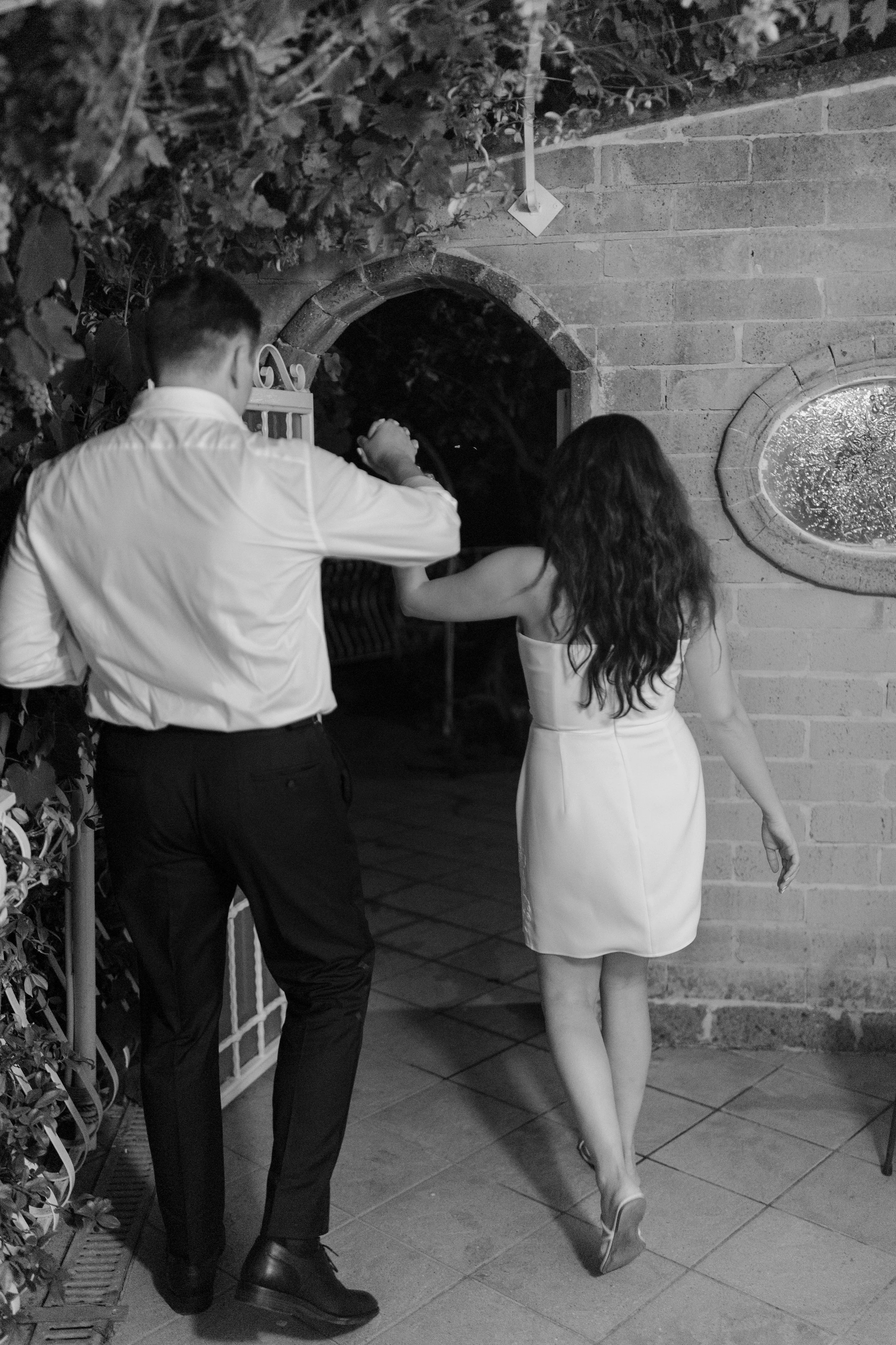 A man and woman are entering a building through a door. The man wears a white shirt and dark pants, and the woman is in a white dress. The scene appears to be at night.